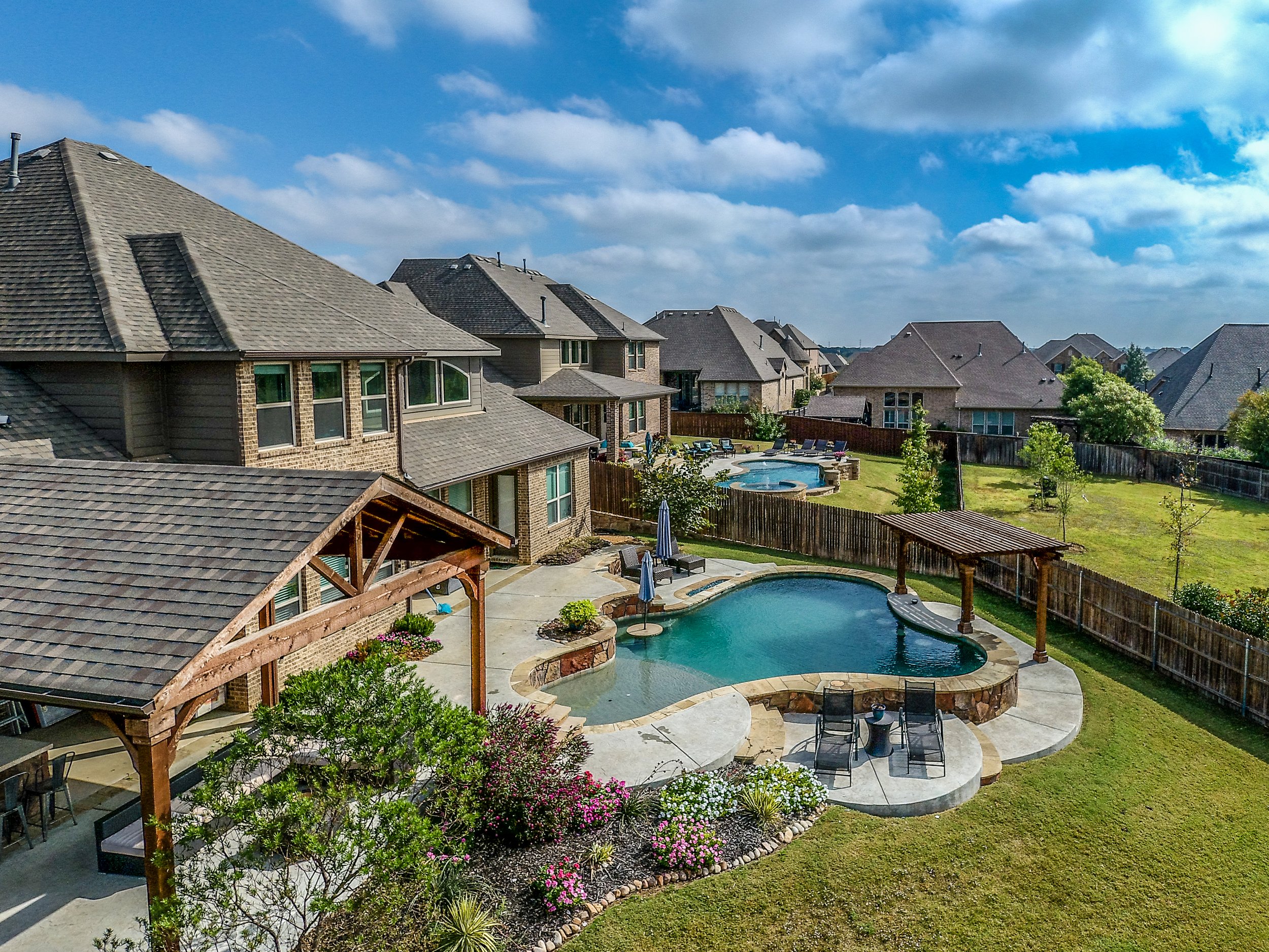 A suburban backyard featuring a swimming pool with a surrounding patio, lounge chairs, and a pergola, with a fenced grassy area and neighboring houses in the background under a partly cloudy sky.