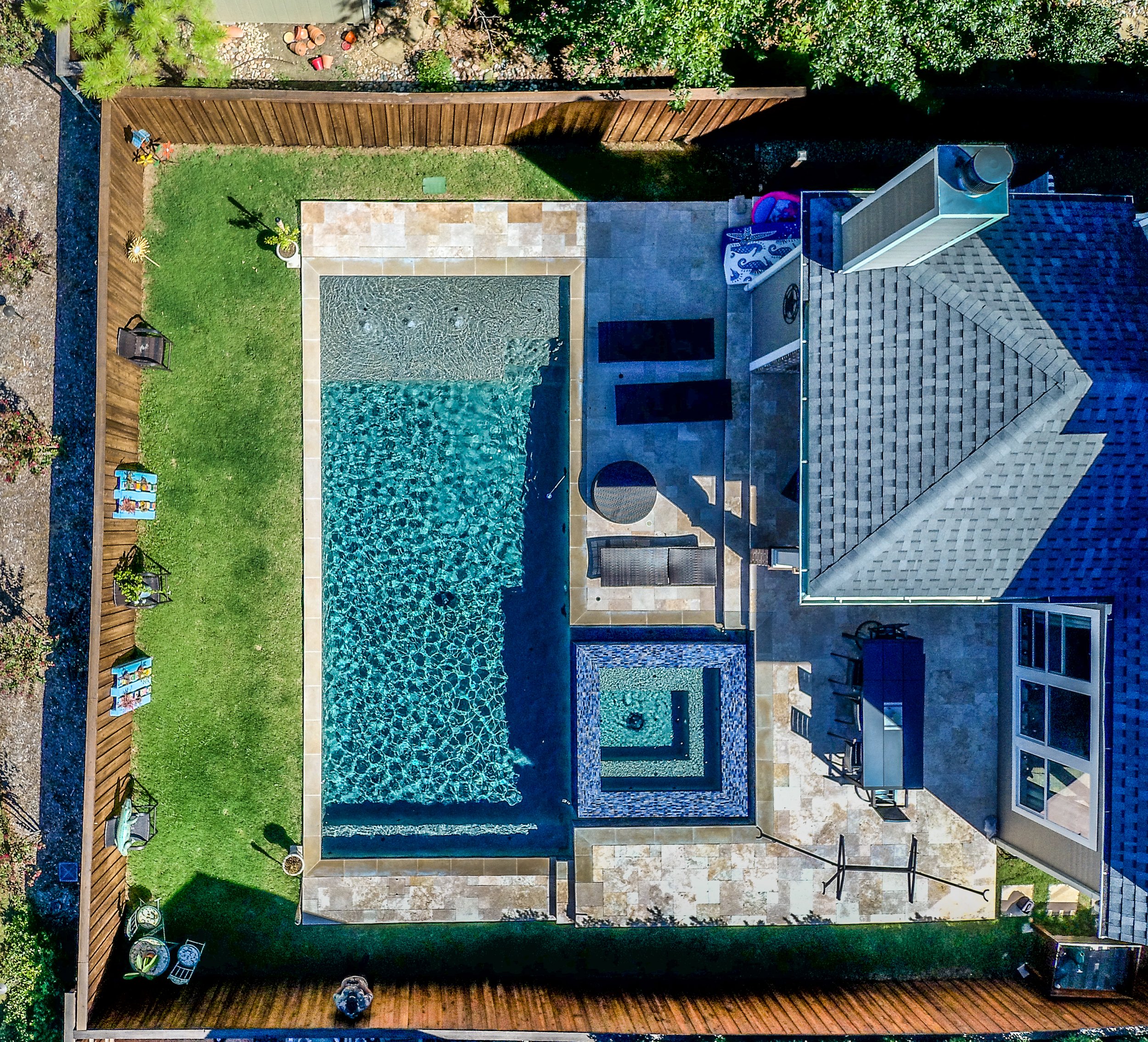 Aerial view of a backyard with a swimming pool, hot tub, patio, lawn, and wooden fence.