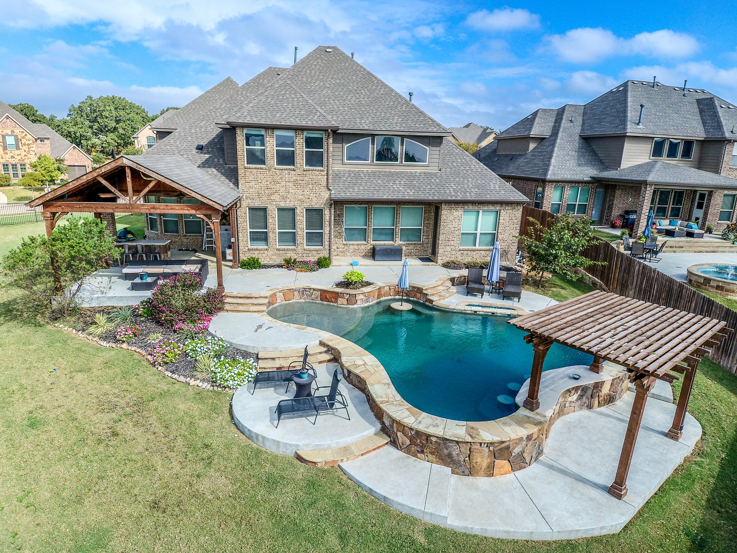 A backyard with a swimming pool, two pergolas, outdoor furniture, and a landscaped garden alongside a large brick house.