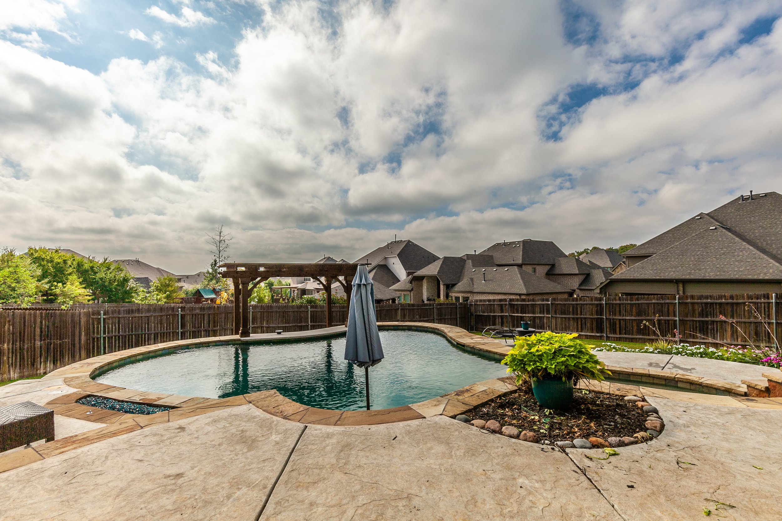 Backyard with a kidney-shaped swimming pool, a closed patio umbrella, a wooden fence, and neighboring houses under a partly cloudy sky.