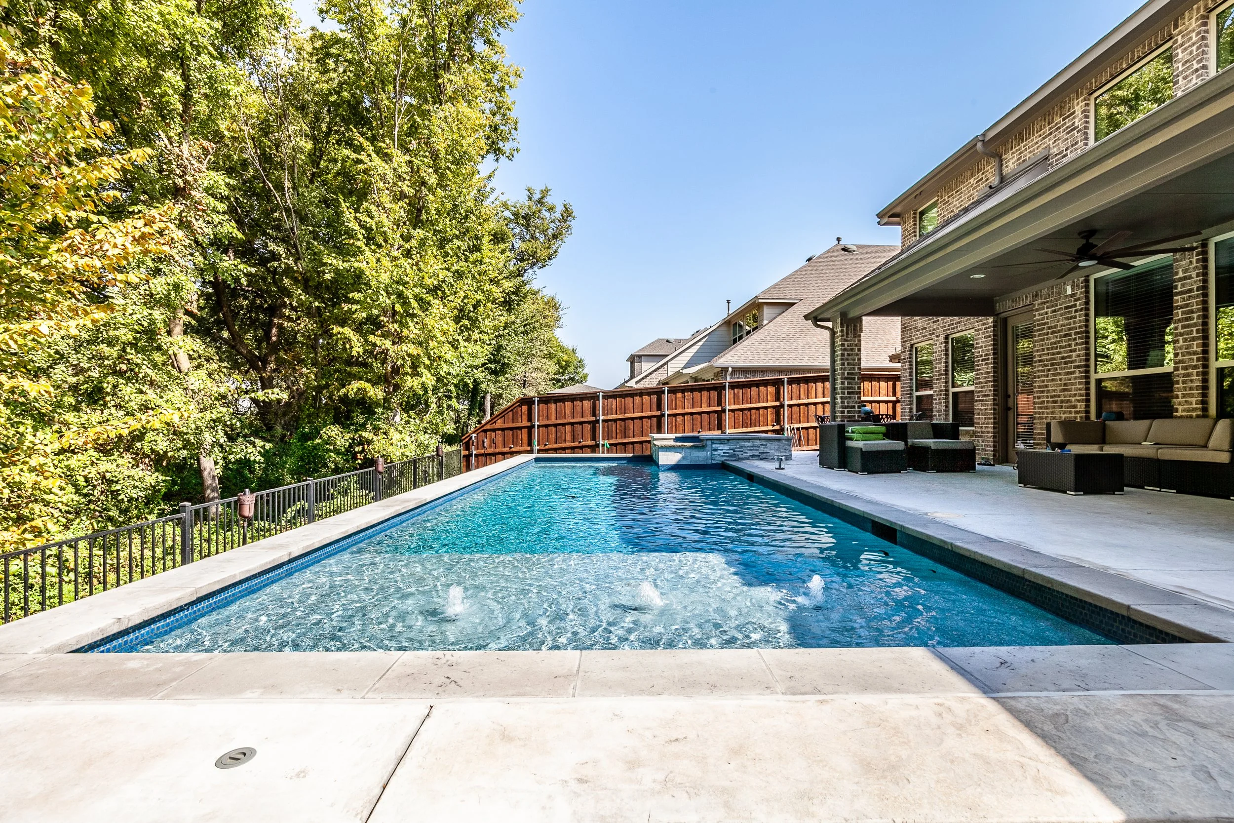 Backyard with a rectangular swimming pool, outdoor seating along a covered patio, green trees on the left, and neighboring houses in the background under a clear blue sky.