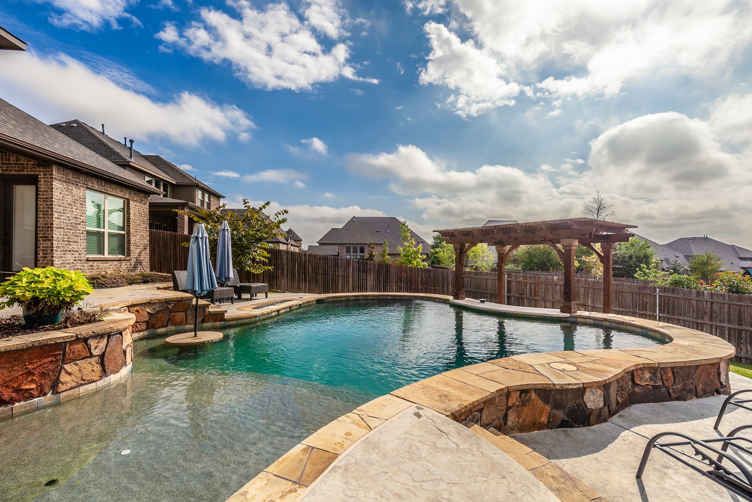 Backyard with a swimming pool, stone patio, two patio umbrellas, lounge chairs, a wooden pergola, wooden fence, and neighboring houses under a partly cloudy sky.
