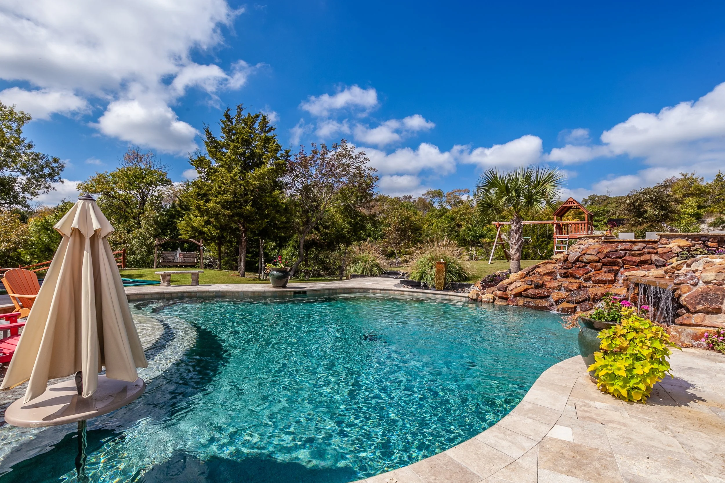Swimming pool in a backyard with trees, a slide, a waterfall feature, chairs, umbrellas, and a blue sky with clouds.