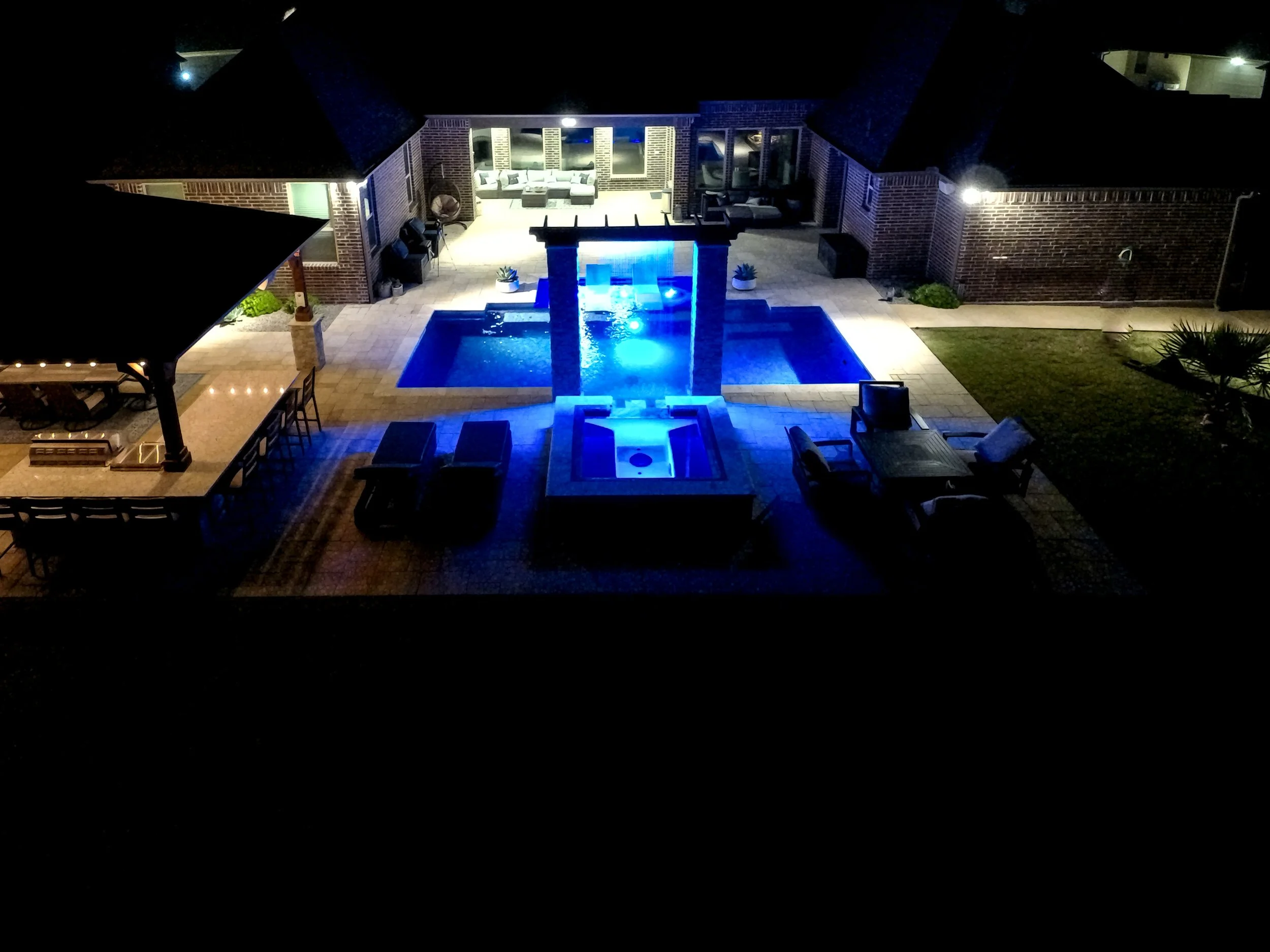 Night view of a backyard with a lit swimming pool, outdoor seating, and a covered patio area.