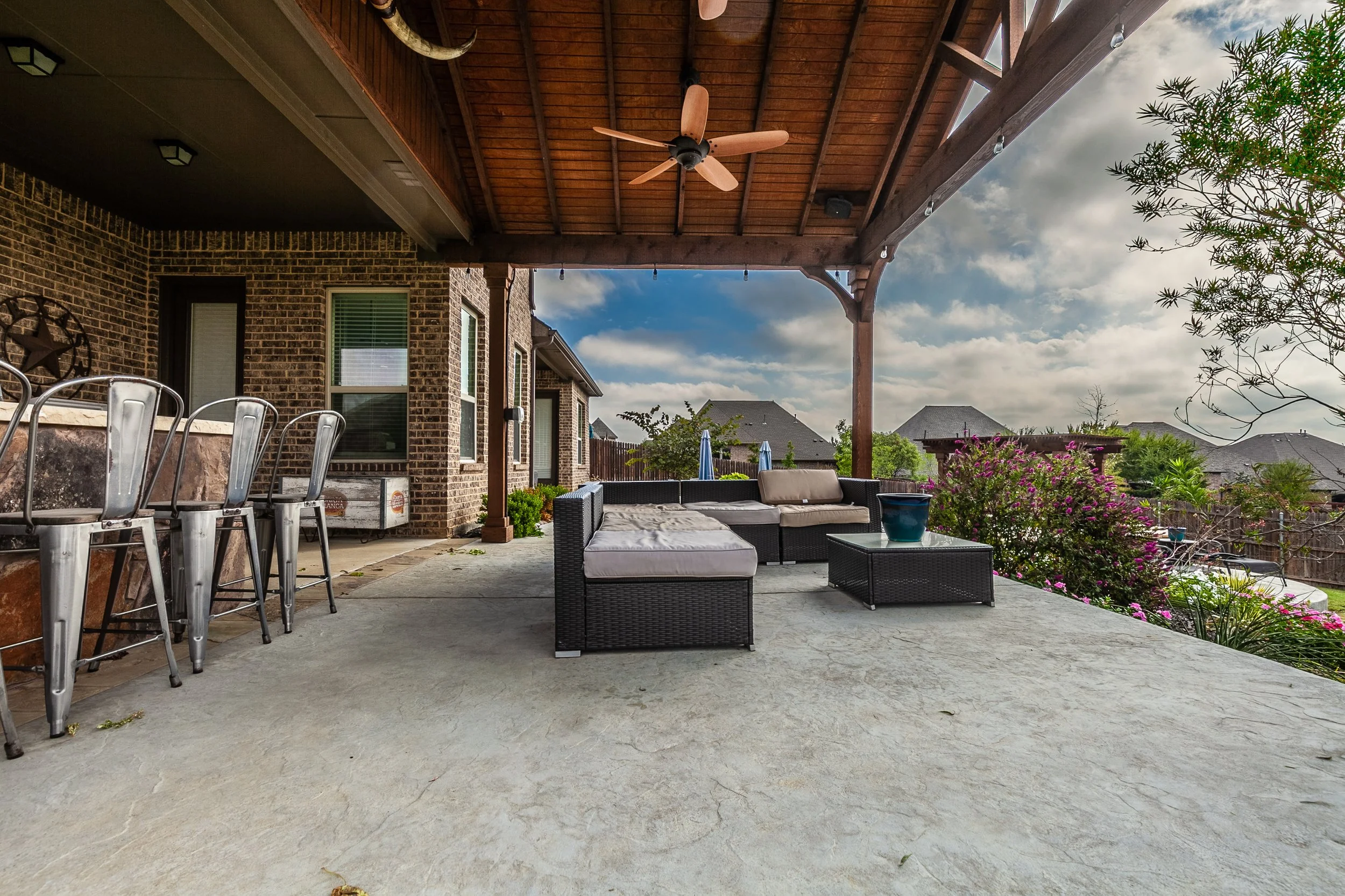 Covered backyard patio with outdoor seating, a brick house wall, green shrubs, pink flowering plants, and a cloudy sky.
