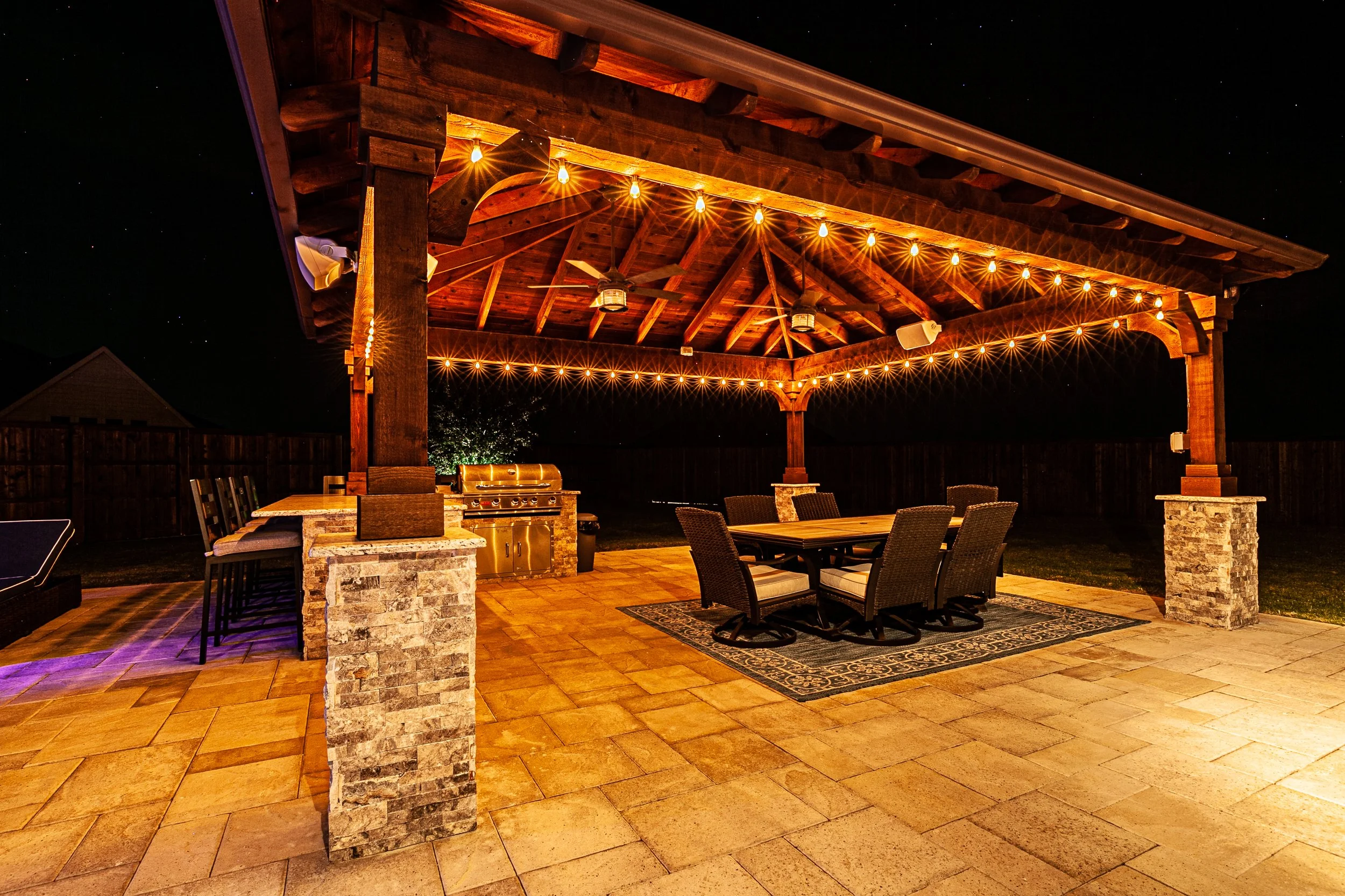Nighttime outdoor patio with wooden pergola, string lights, dining table with six chairs, corner barbecue grill, and stone pillars, illuminated softly.