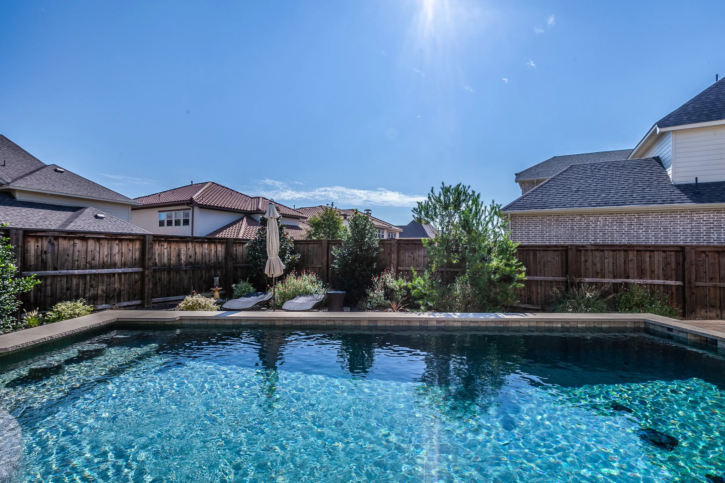 Backyard with a swimming pool, wooden fence, outdoor lounge chairs, a closed umbrella, trees, and neighboring houses under a bright sunny sky.