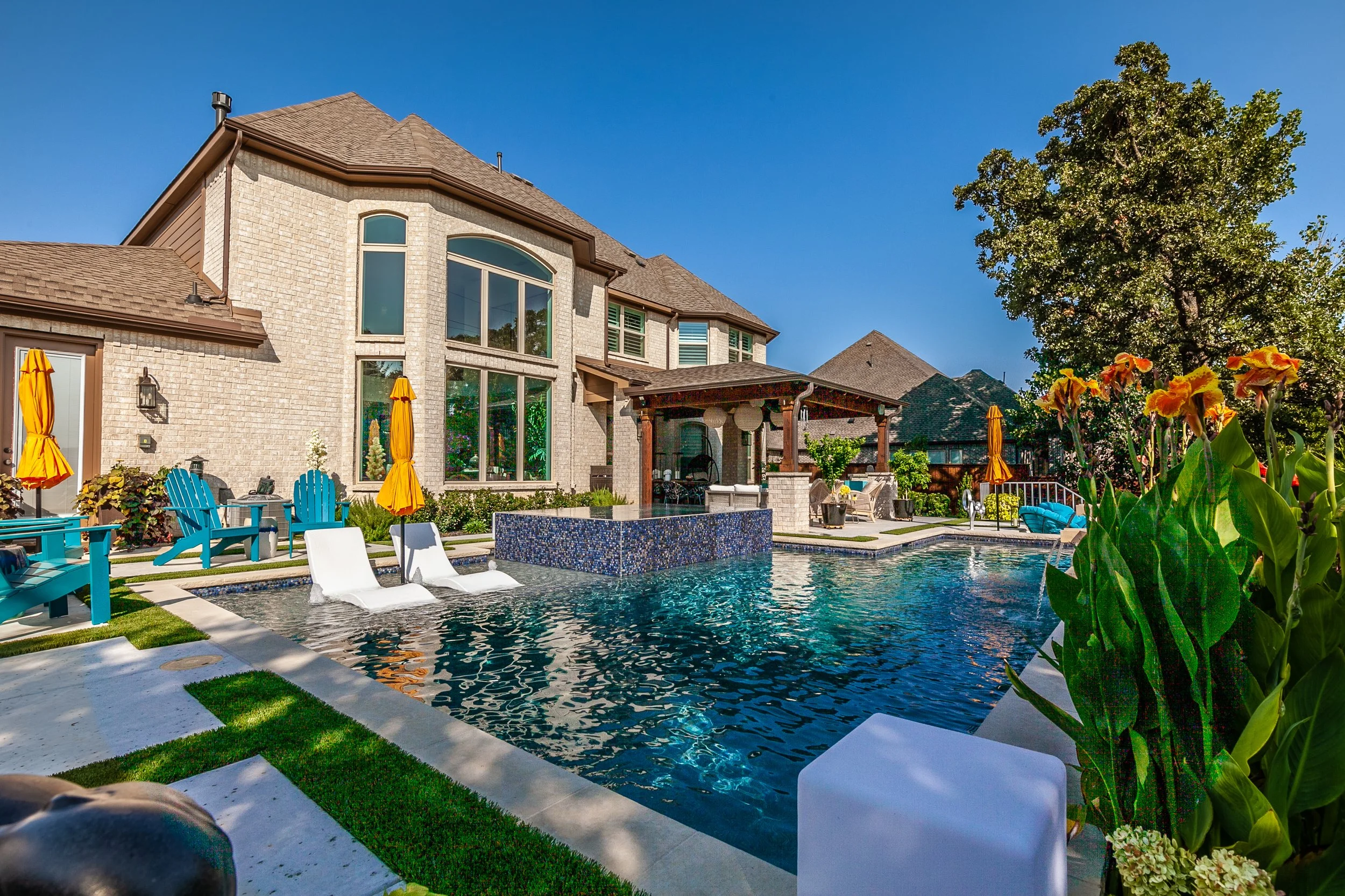 Backyard with a modern house, swimming pool, orange umbrellas, blue Adirondack chairs, and lush landscaping under a clear blue sky.