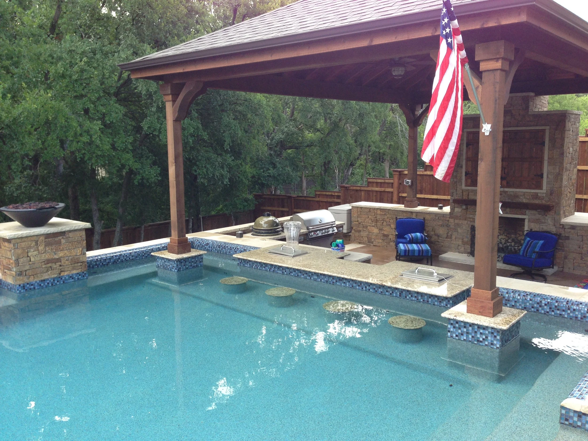 Backyard swimming pool with a covered patio area, barbecue grill, outdoor seating, and an American flag hanging from the patio structure. Surrounding trees are visible in the background.
