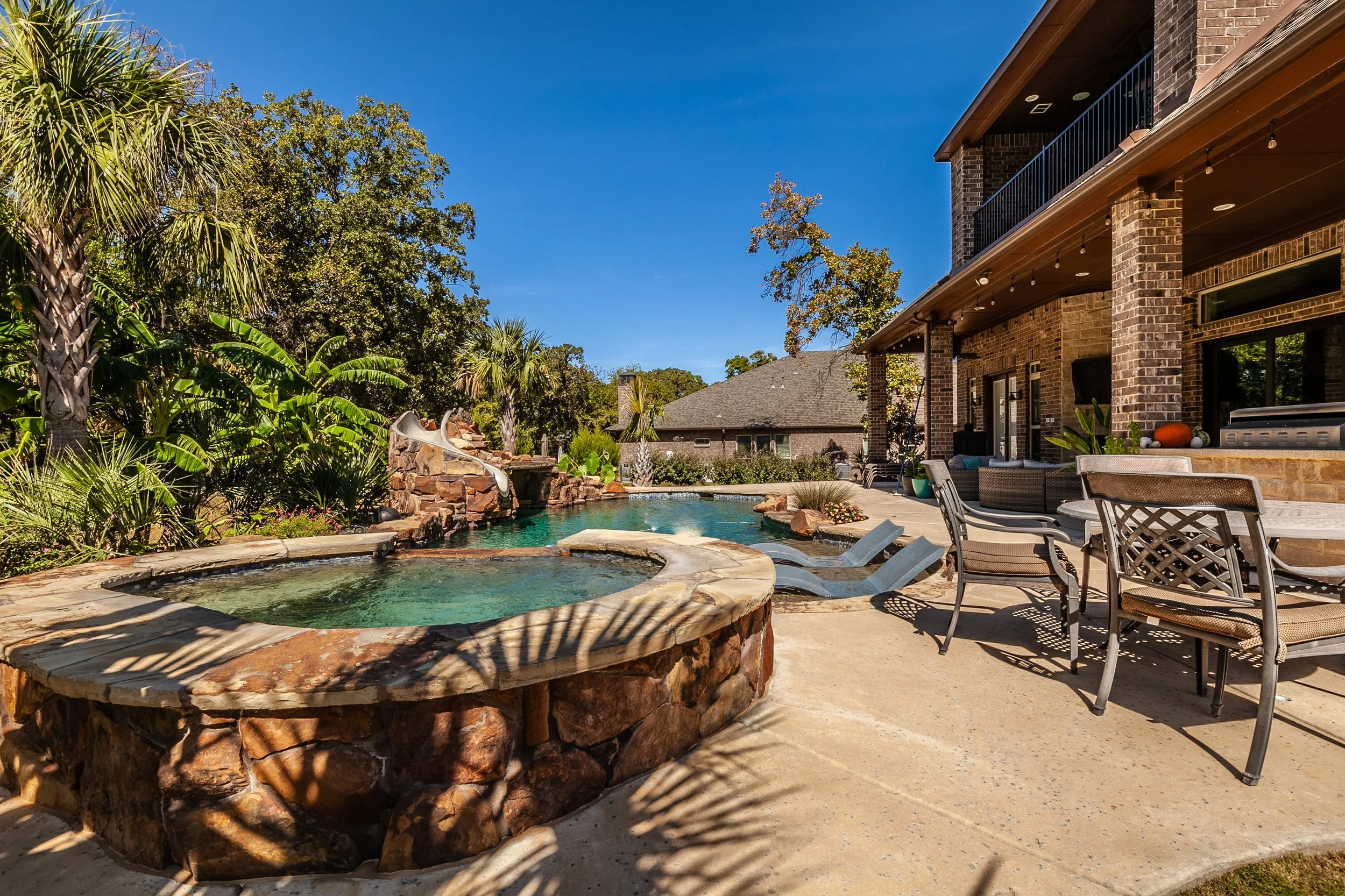 Backyard with a swimming pool and hot tub, surrounded by tropical and leafy trees, patio furniture, and a brick house with a covered porch.