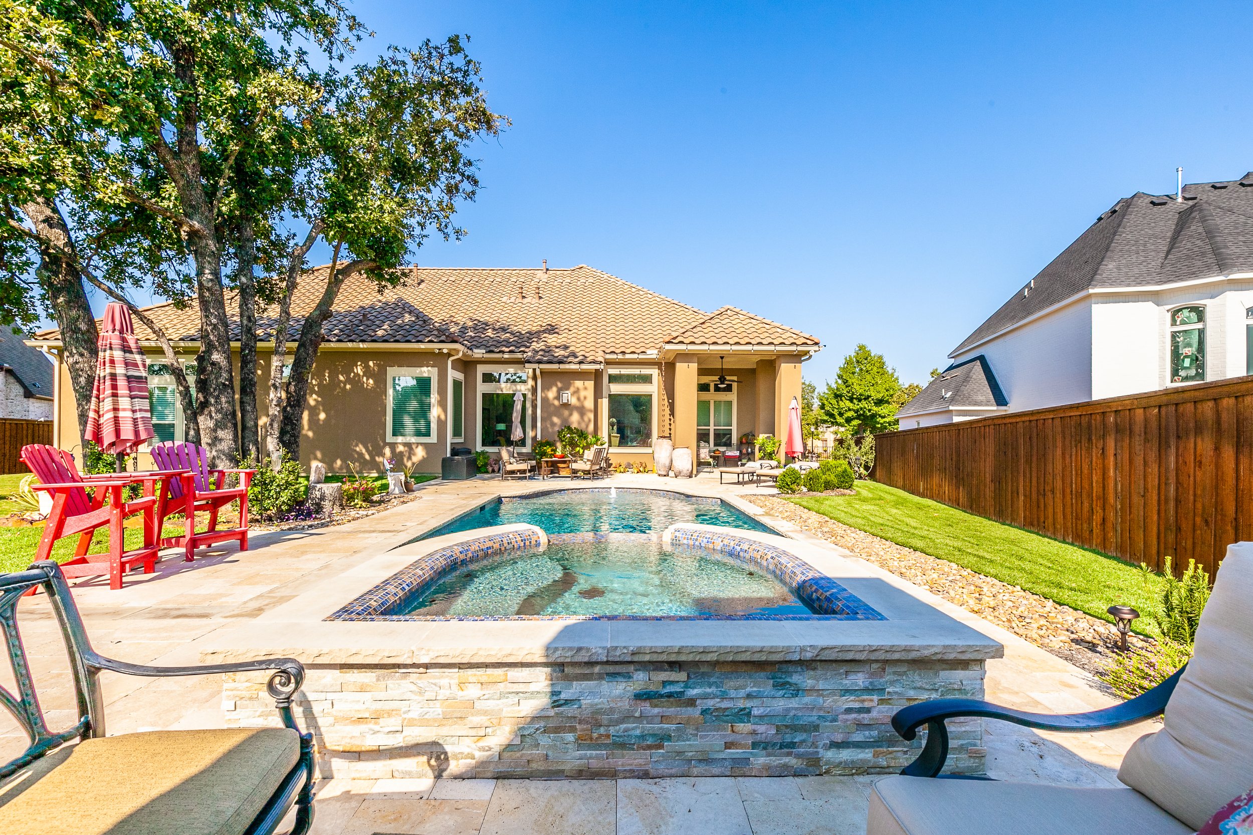 Backyard with a swimming pool, patio furniture, trees, and neighboring houses under a clear blue sky.