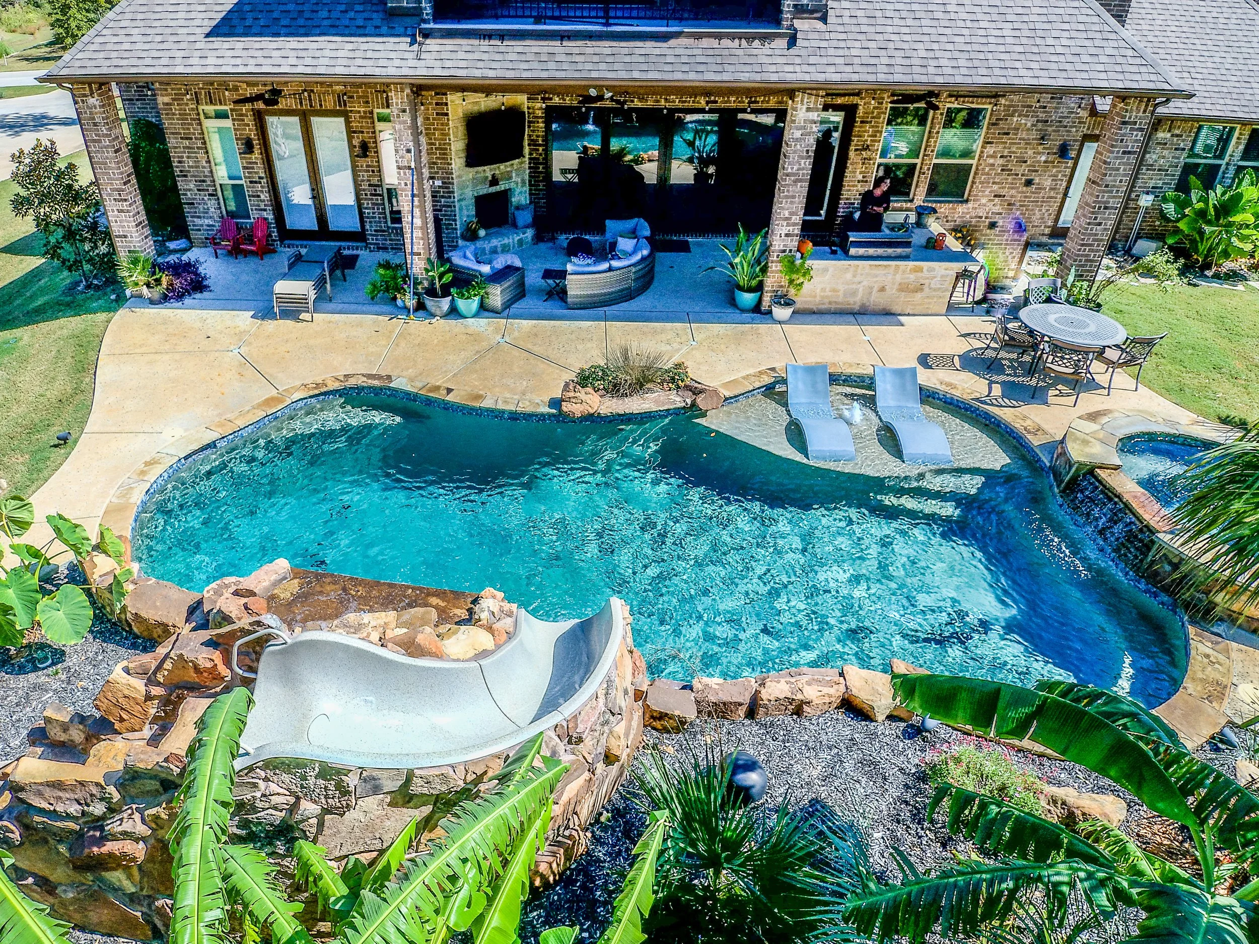 A backyard with a swimming pool, a slide, outdoor furniture, and a brick house with large windows and a patio area.
