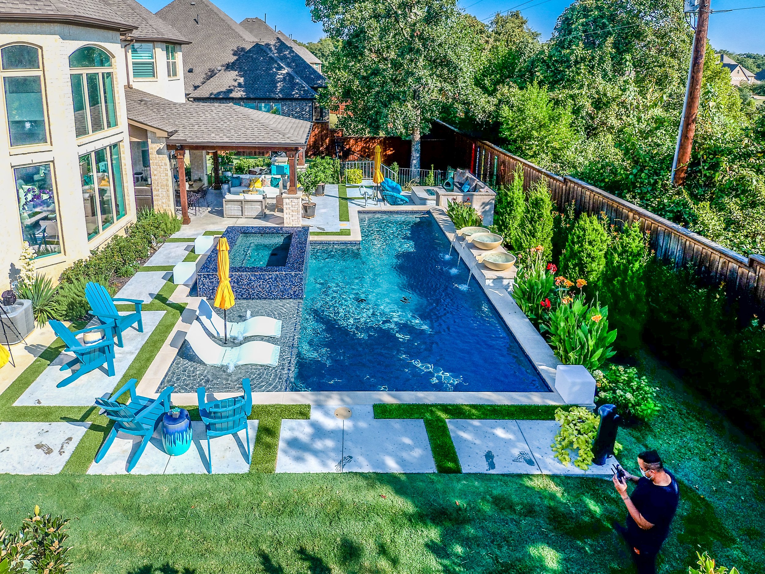 A backyard with a swimming pool surrounded by chairs and umbrellas, outdoor furniture under a covered patio, lush greenery, and a man wearing a mask standing on the grass looking at his phone.