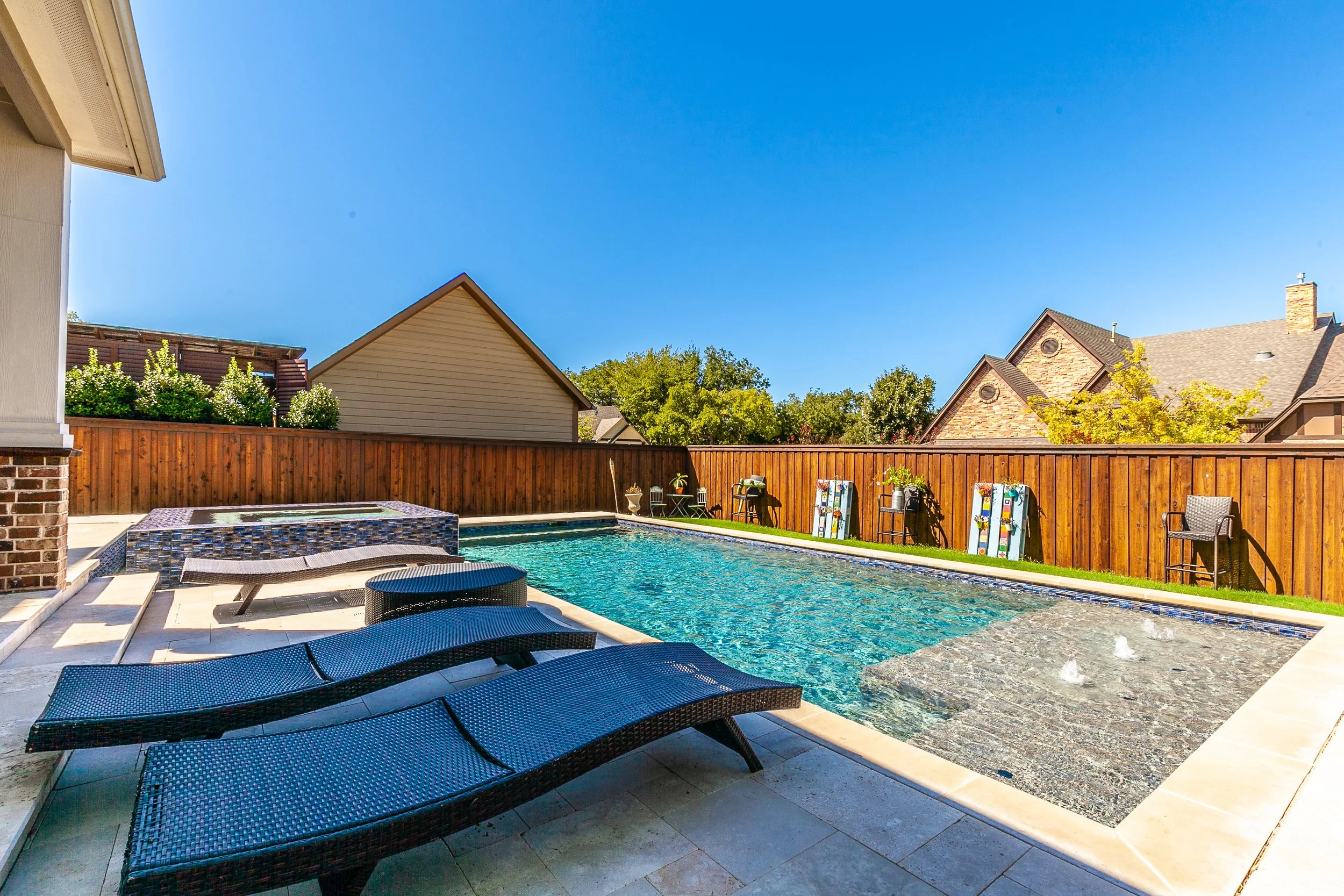 Backyard swimming pool with lounge chairs, a small fountain feature, and a wooden fence, in a sunny residential area.