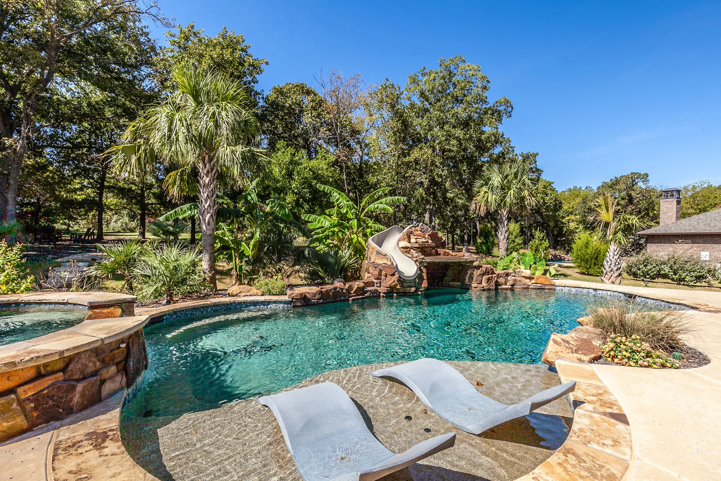 A backyard swimming pool with a rock water slide, surrounded by lush green trees and plants, and two white lounge chairs on the pool deck.