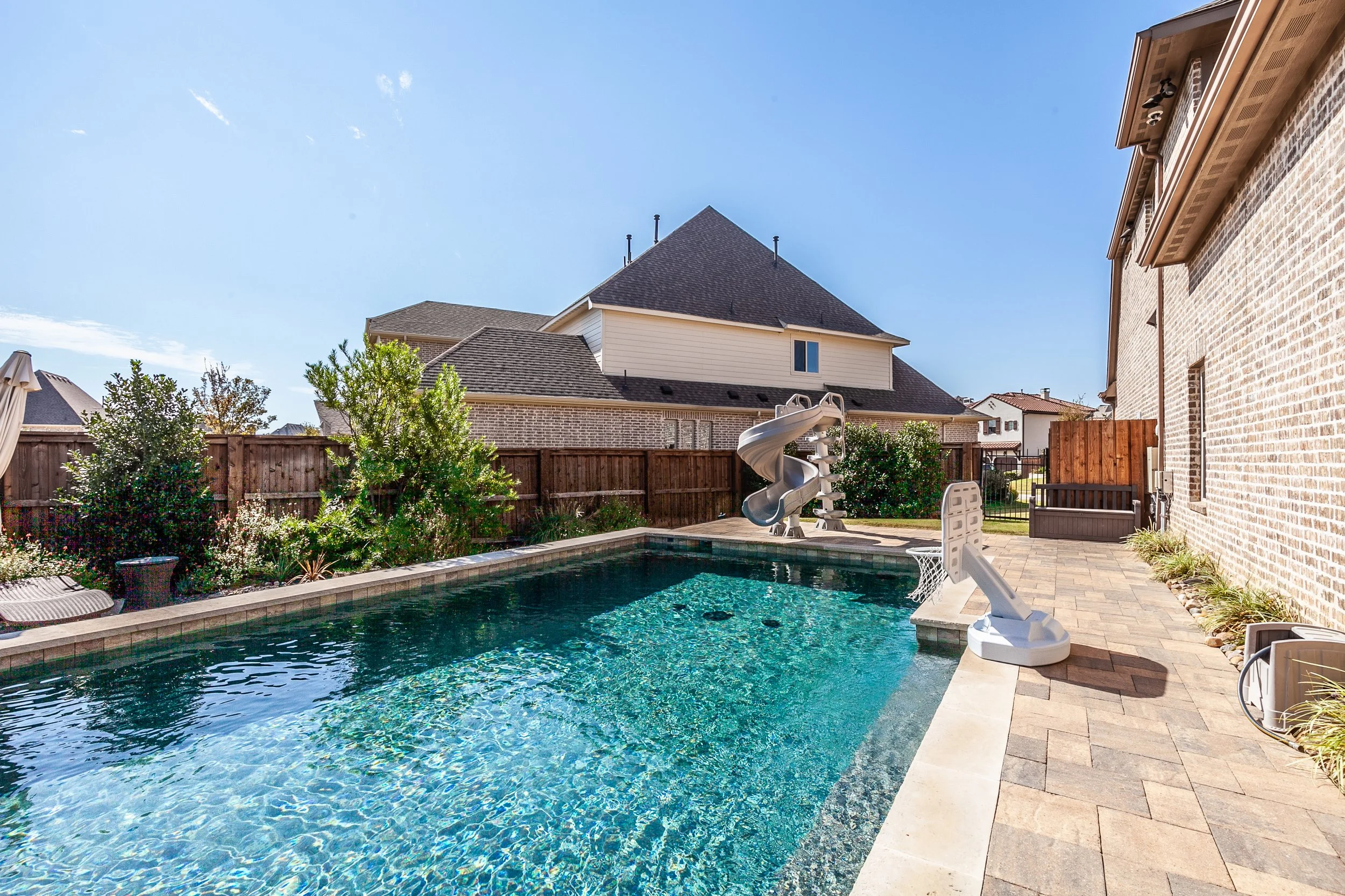 A backyard swimming pool with a water slide and small basketball hoop, surrounded by a paved patio and a garden, with a brick and siding house and a wooden fence in the background under a clear blue sky.