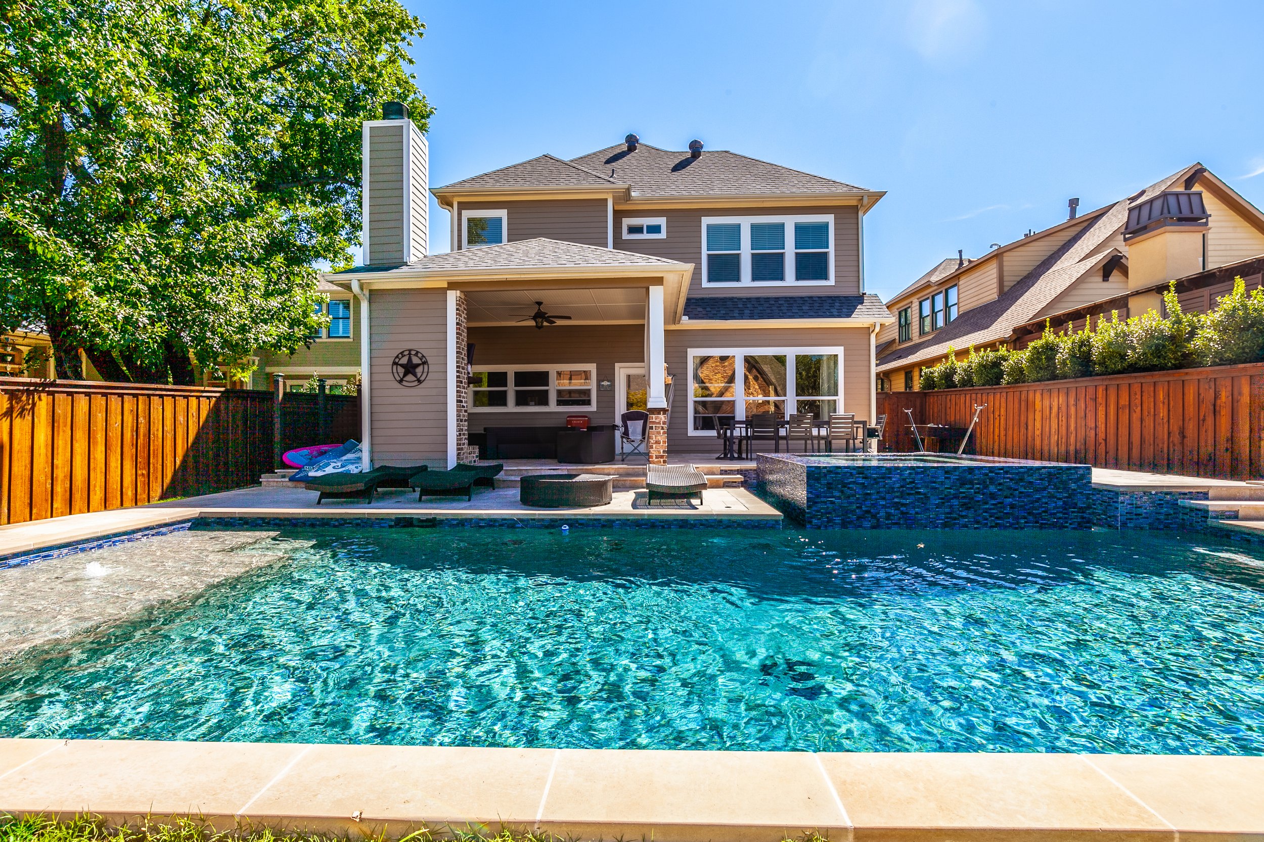 Backyard with a swimming pool, outdoor seating, and a two-story house with gray siding and large windows under a clear blue sky.