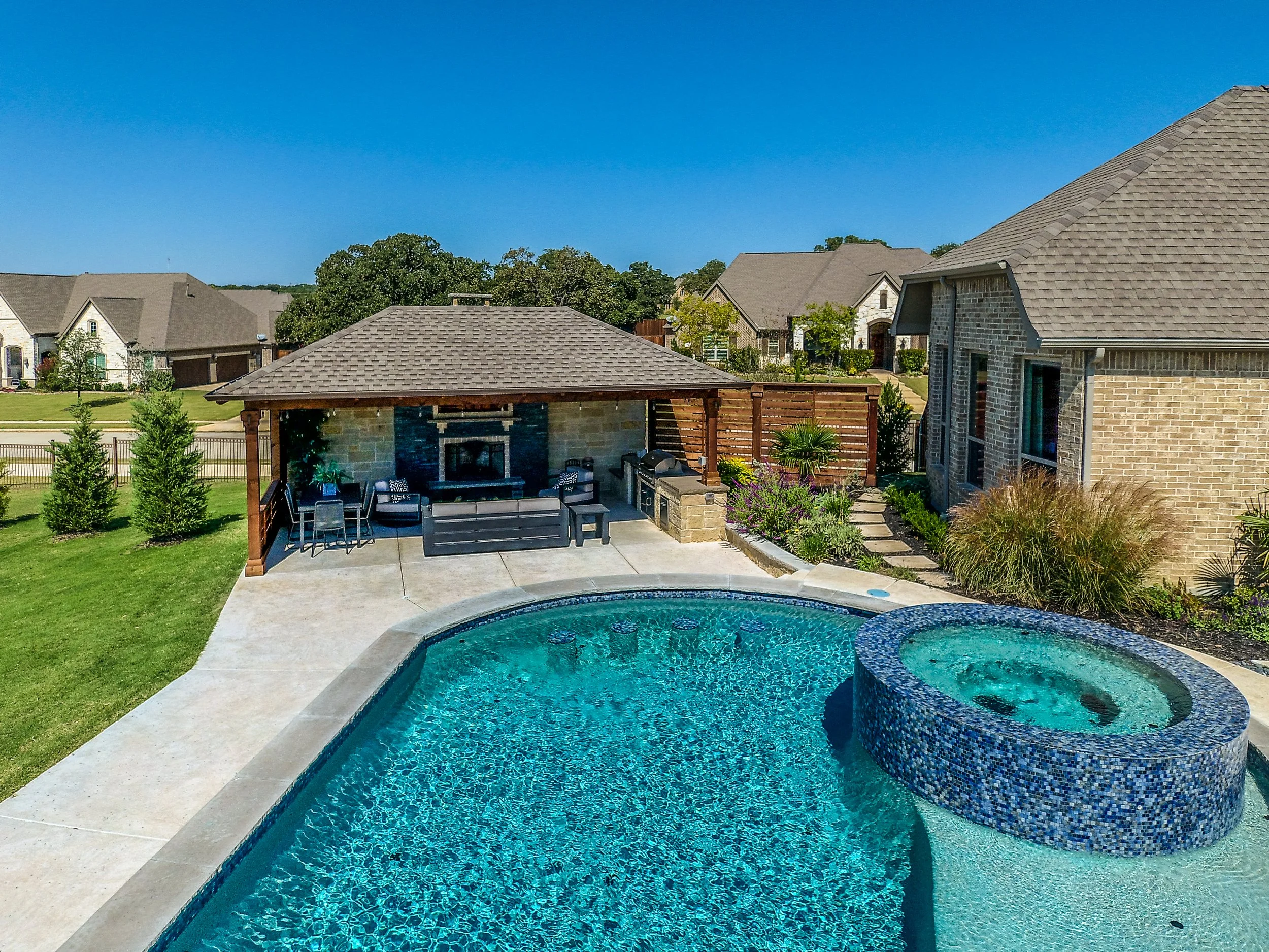 A backyard pool with a hot tub, outdoor seating area under a covered patio, surrounding landscaping, and neighboring houses in the background.