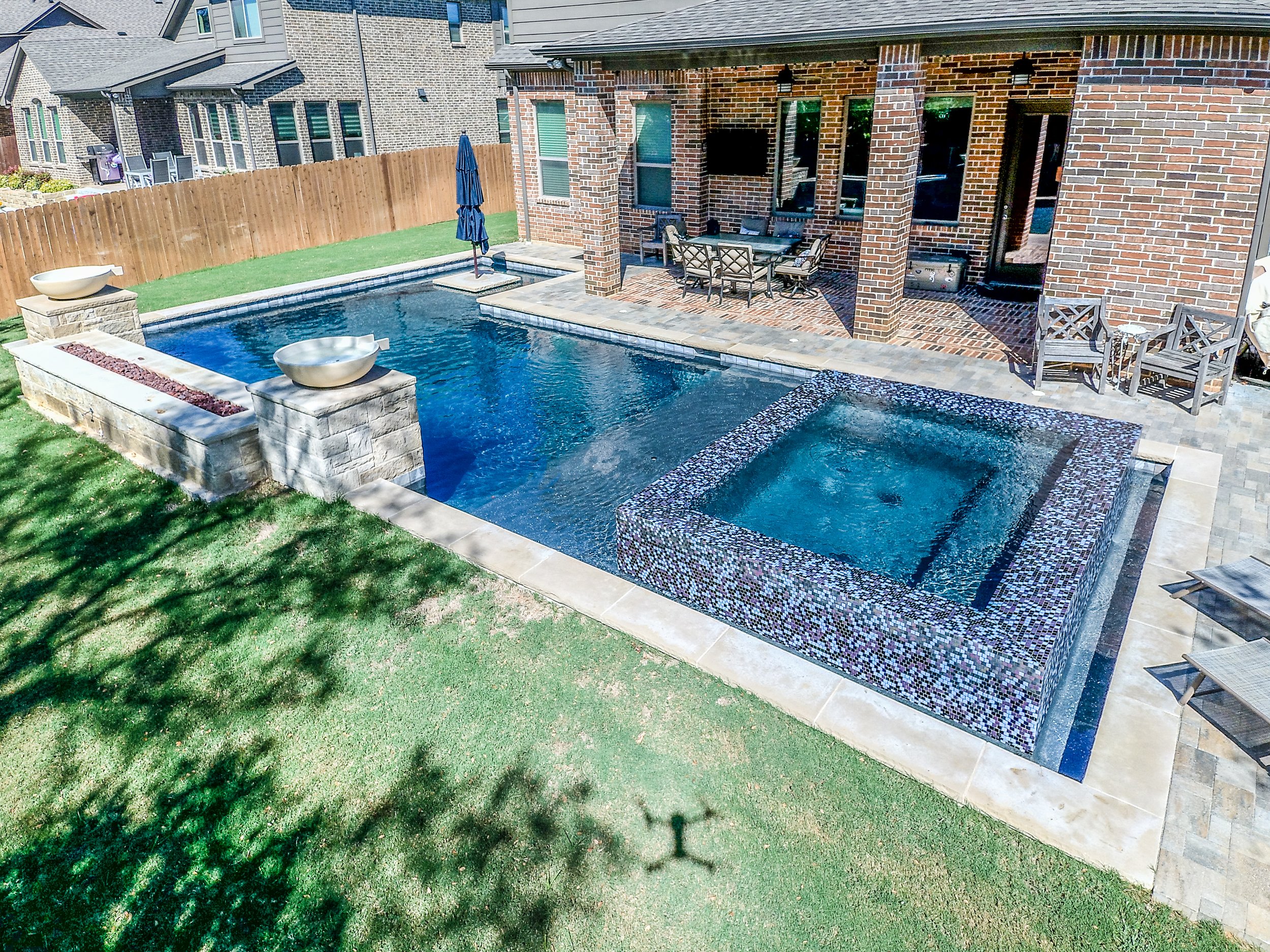 Backyard with a swimming pool featuring a raised hot tub, patio furniture, a brick house with a covered porch, and a wooden fence.