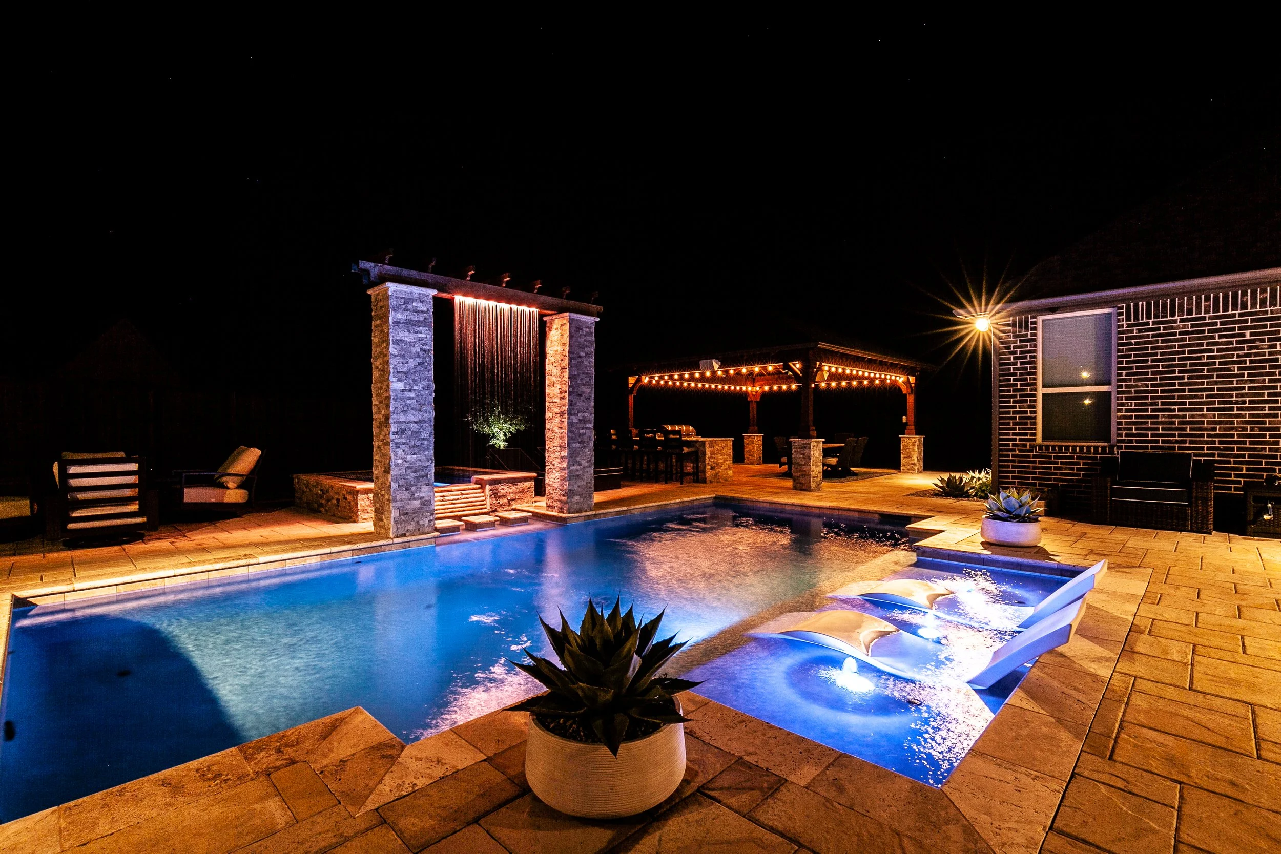 Nighttime view of a lit outdoor swimming pool with a built-in hot tub, surrounded by patio furniture, potted plants, string lights, and a gazebo with seating.