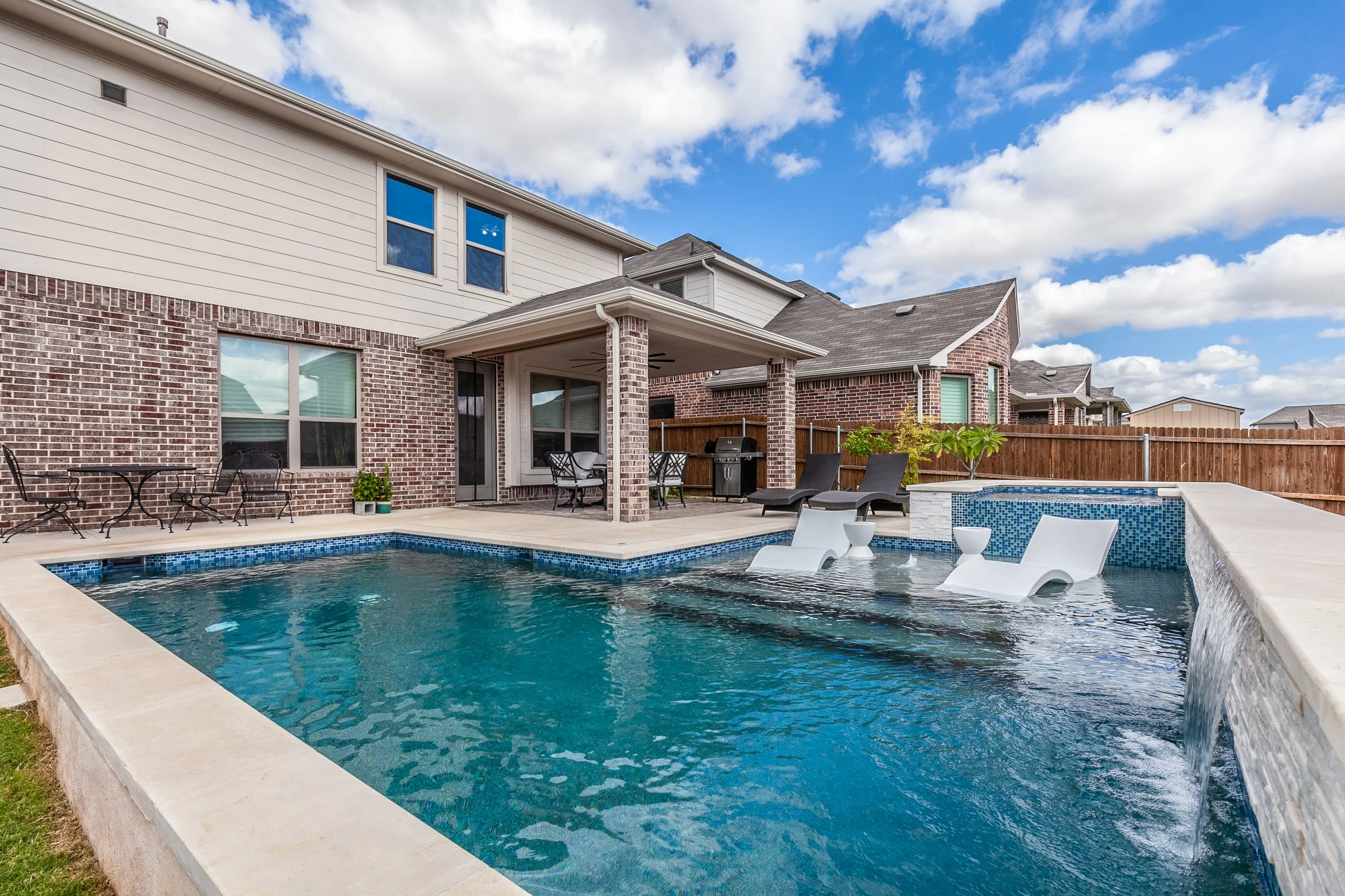 Backyard with a swimming pool, patio furniture, and a covered patio area, surrounded by a brown wooden fence, under a partly cloudy sky.