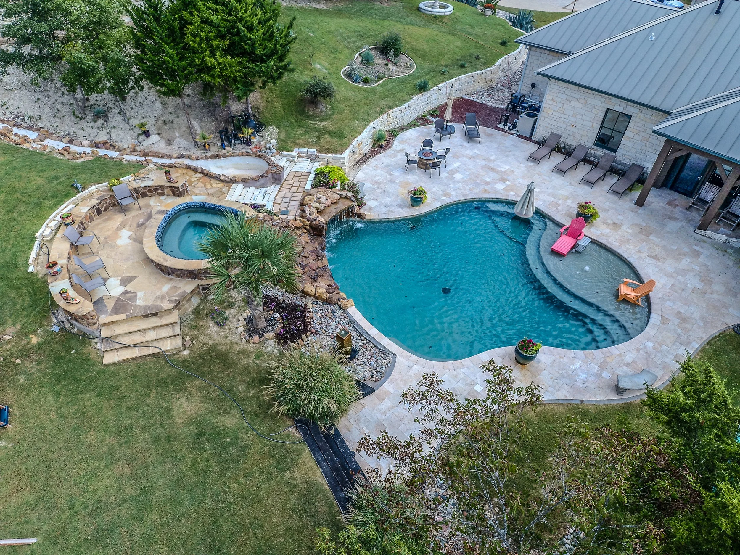 Aerial view of a backyard with a large kidney-shaped swimming pool, a hot tub, and various outdoor furniture including lounge chairs, tables, and umbrellas. The yard is landscaped with grass, trees, and decorative plants, and a stone patio surrounds 