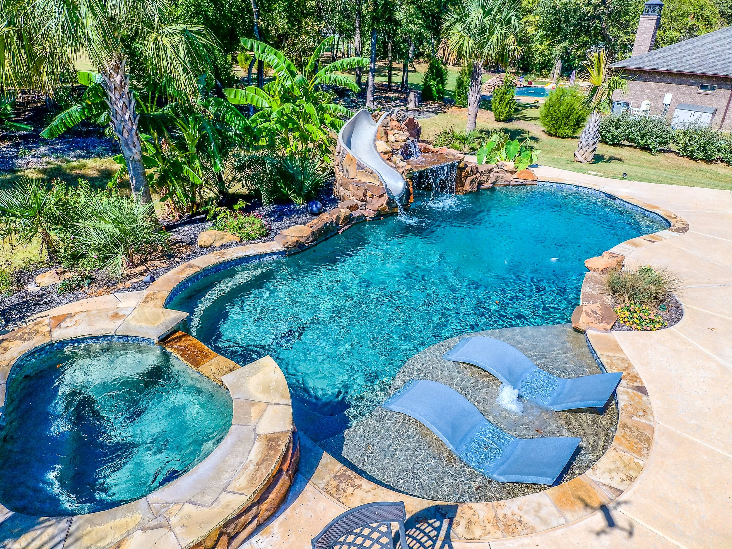 A backyard swimming pool area with a hot tub, a slide, and multiple lounge chairs surrounded by tropical plants and trees.