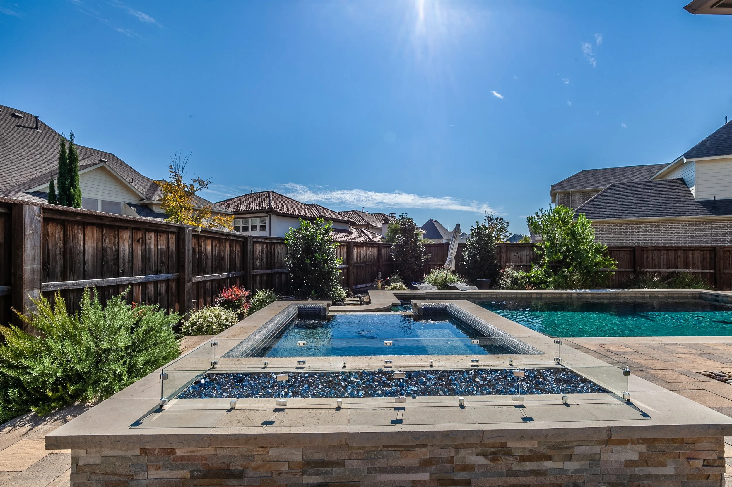 Backyard with a swimming pool and hot tub, surrounded by a wooden fence, trees, and houses under a blue sky.