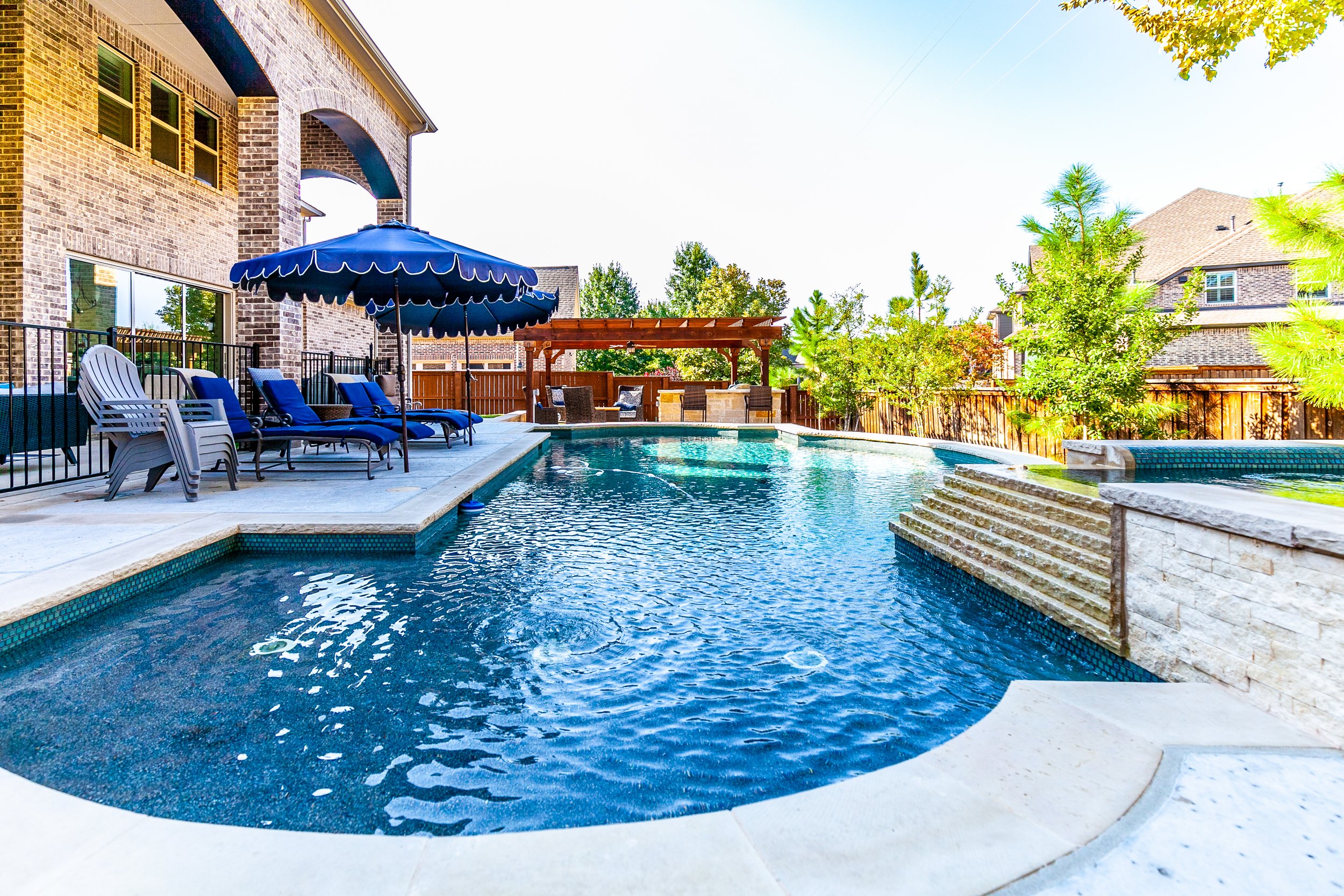 Backyard with a swimming pool, blue lounge chairs, a large blue umbrella, a wooden gazebo with outdoor seating, green trees, and a brick house in the background.