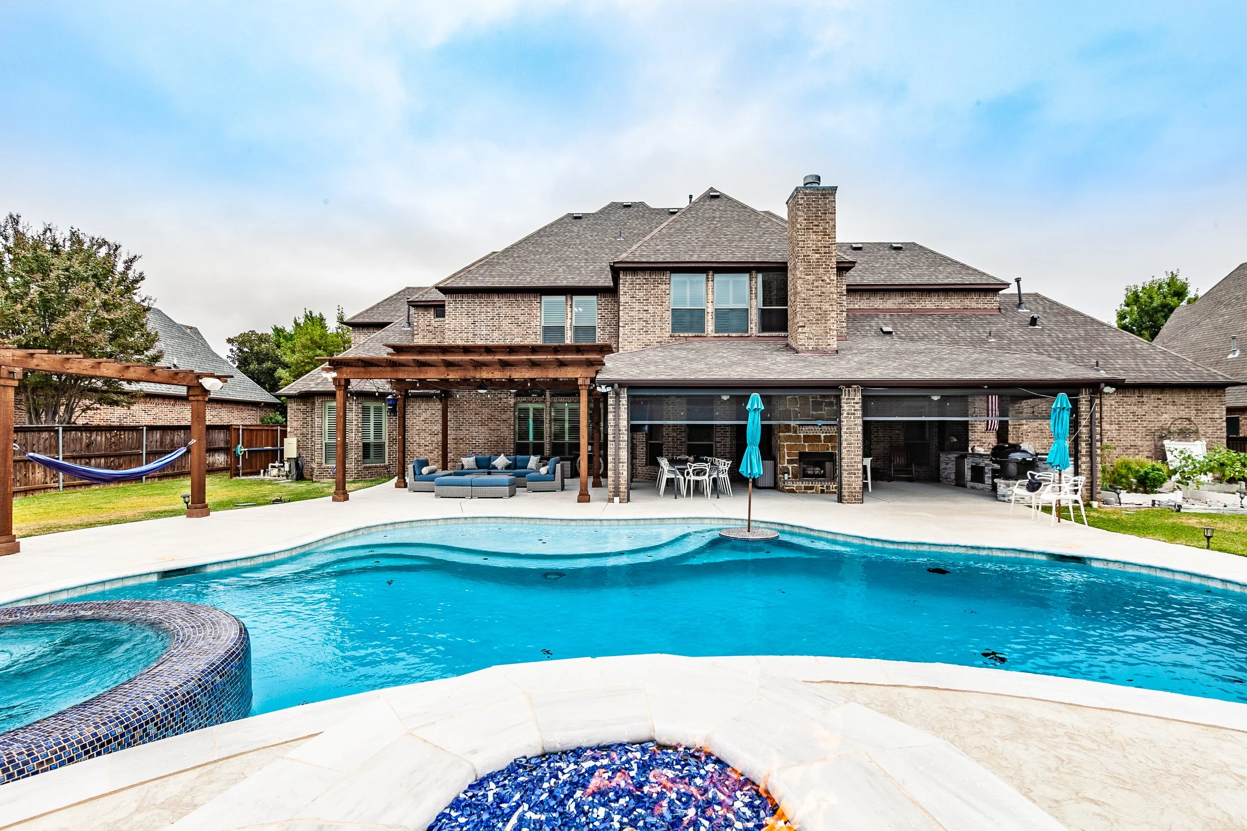 Backyard view of a large brick house with a swimming pool, hot tub, and patio furniture.