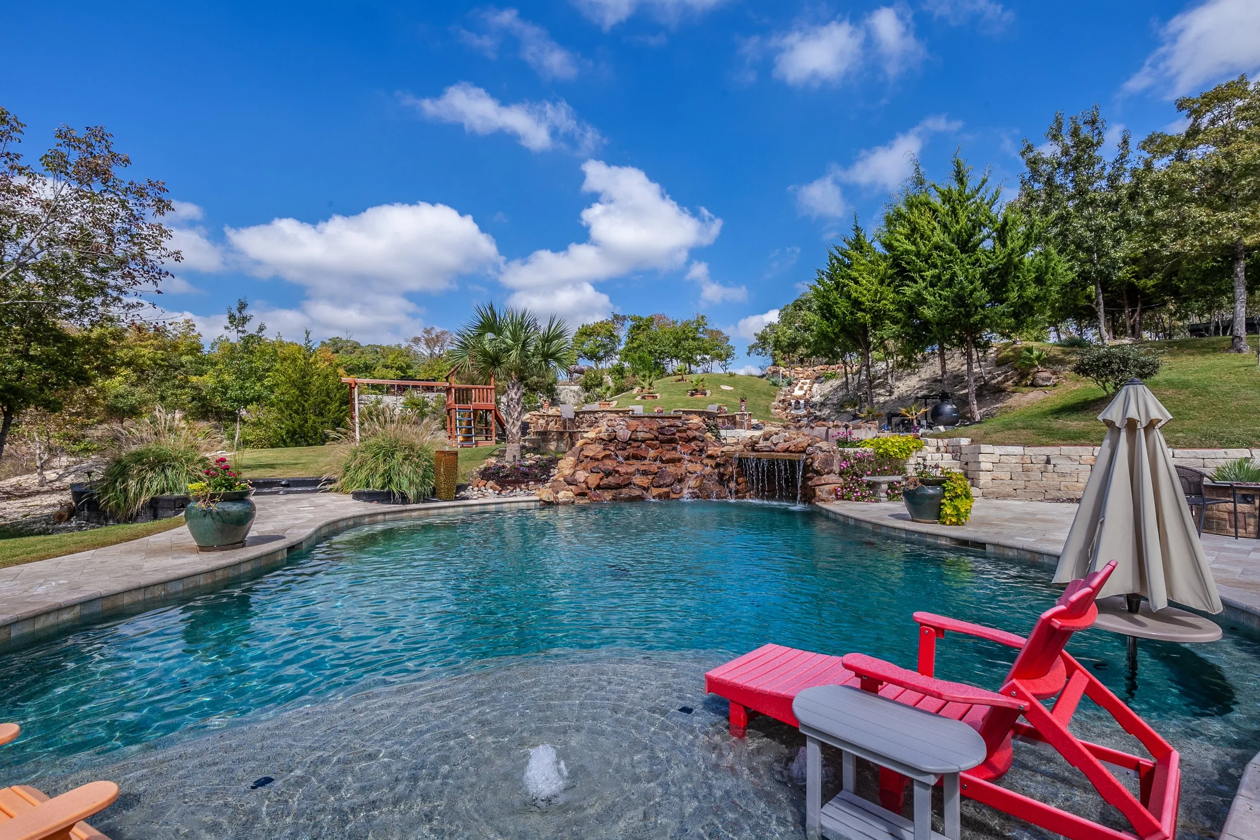 Backyard swimming pool with a waterfall feature, surrounded by potted plants, trees, and outdoor furniture, under a blue sky with scattered clouds.