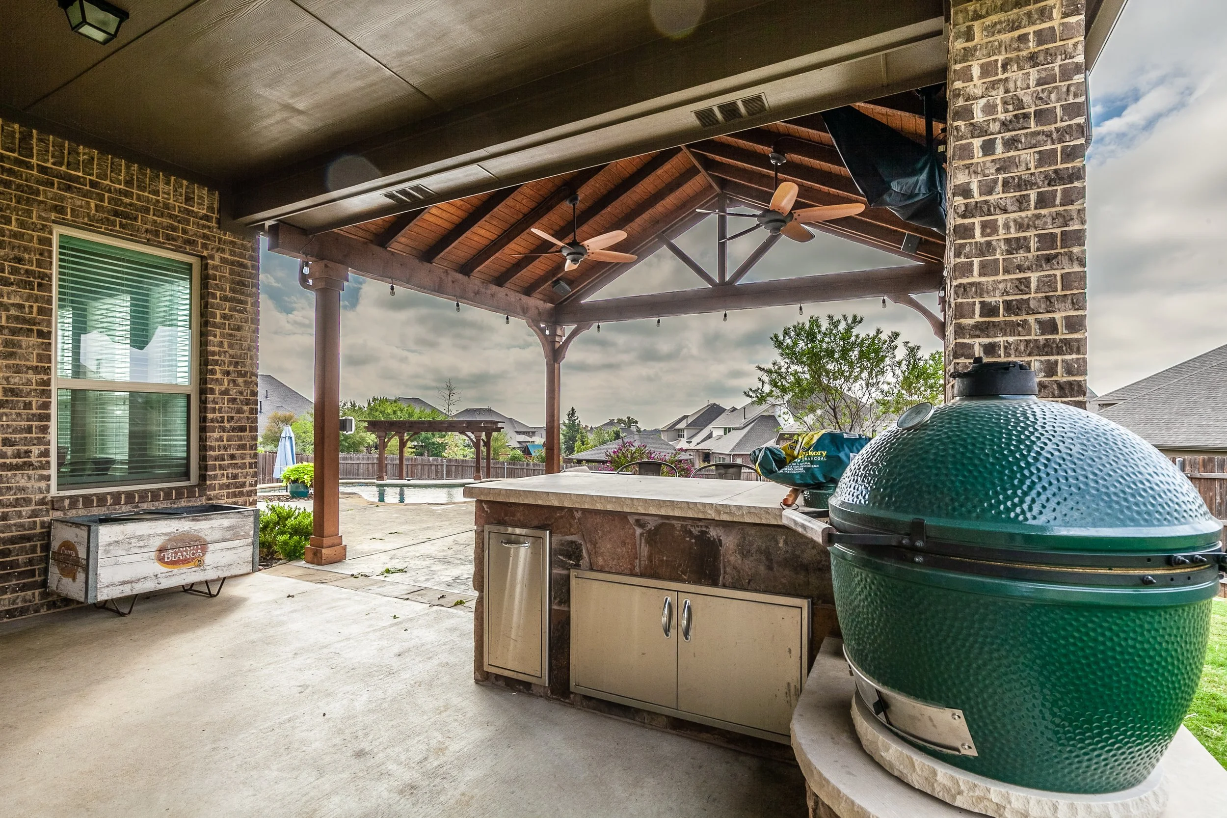 Covered outdoor patio with brick columns, ceiling fans, and a large green smoker. There is a countertop with cabinet storage, a pool area with a gazebo and trees in a backyard, and a nearby window and storage box.