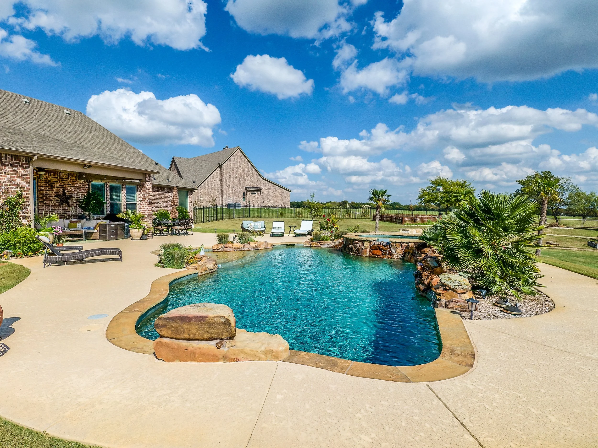 Backyard with a swimming pool, surrounded by loungers, chairs, and outdoor furniture, with a brick house and a clear blue sky with clouds in the background.