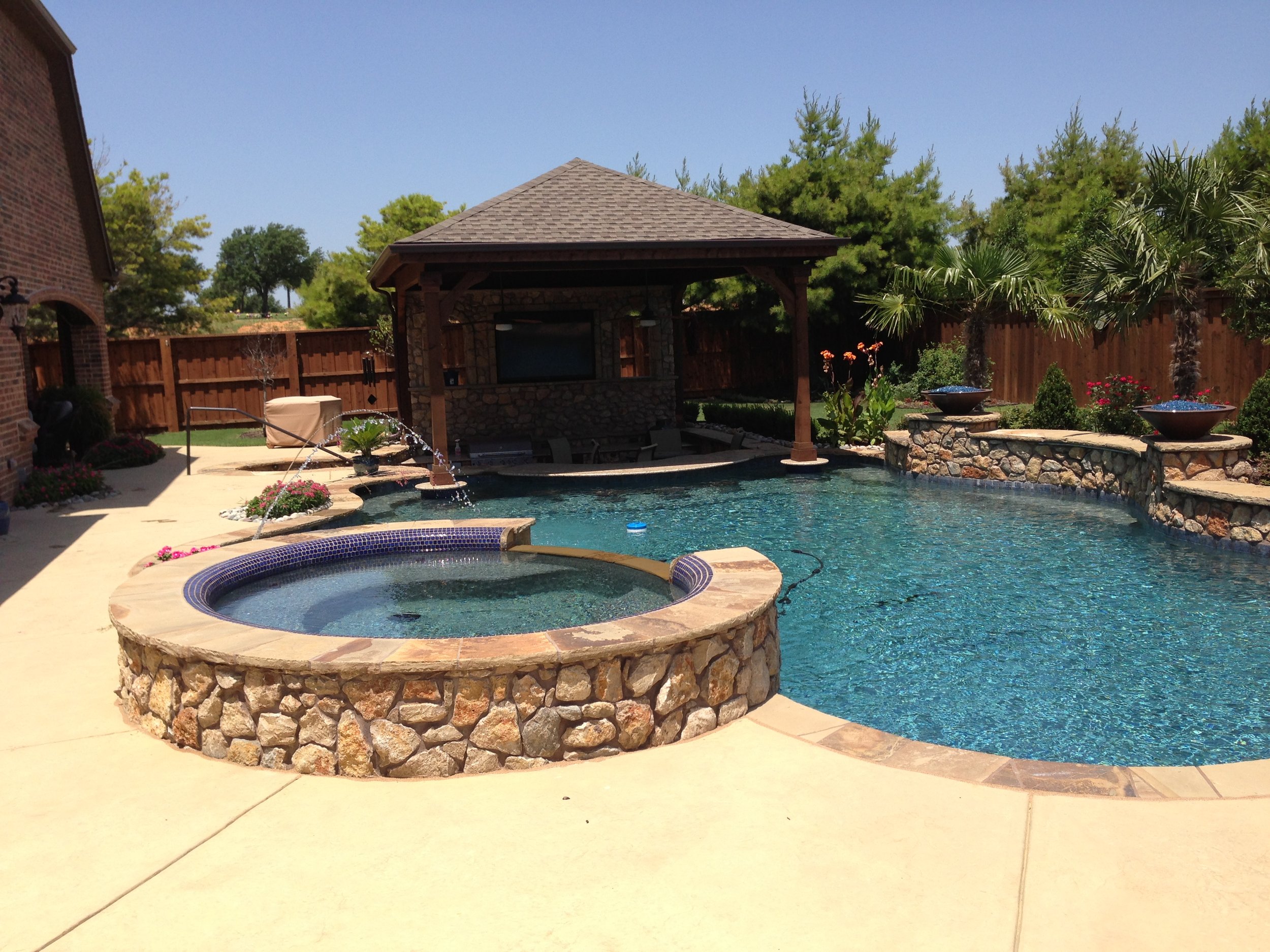 Backyard swimming pool with a hot tub, surrounded by a concrete patio, with a wooden fence, palm trees, and a covered seating area in the background.