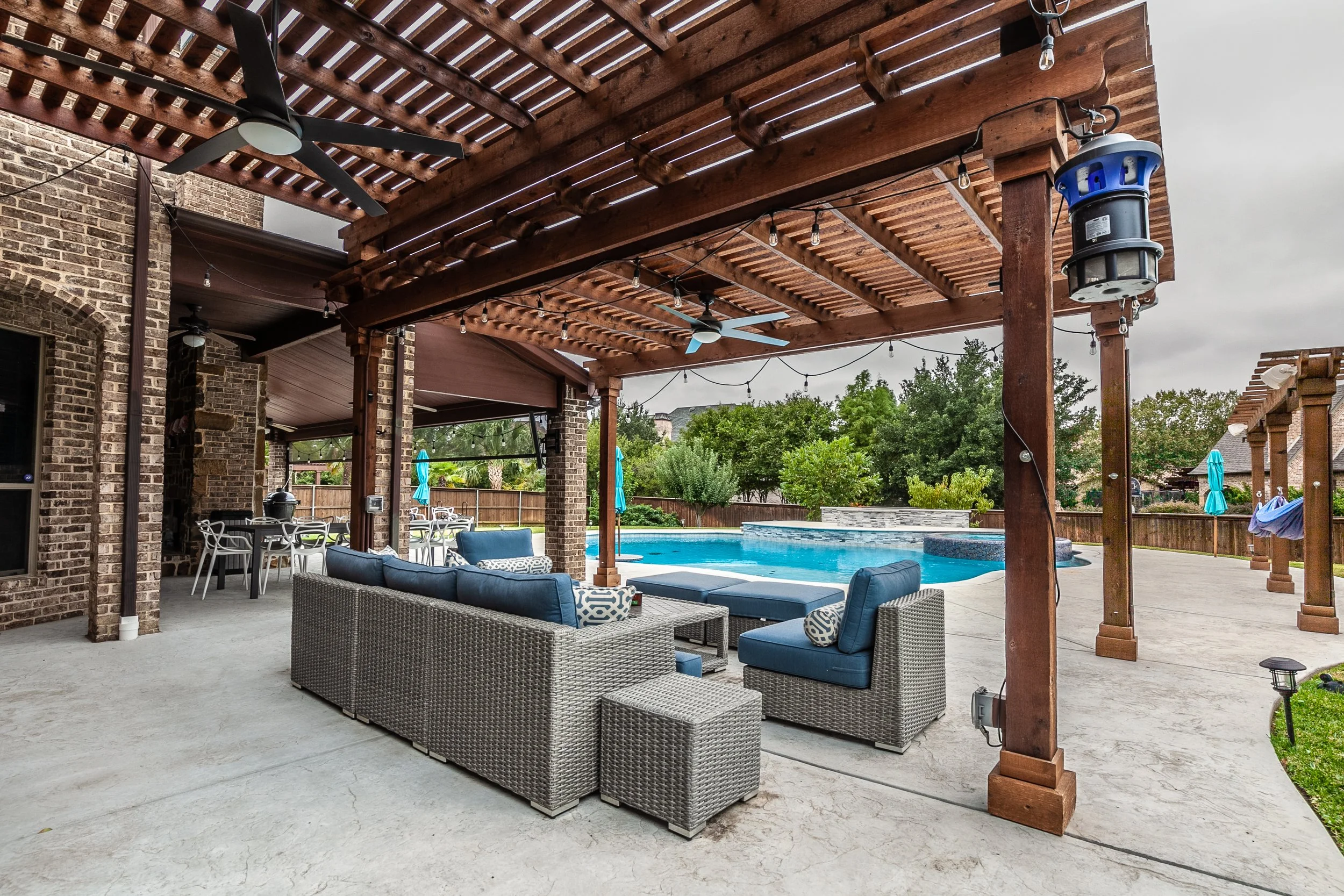 Outdoor backyard patio with a swimming pool, wicker furniture with blue cushions, and a wooden pergola with string lights and ceiling fans under a cloudy sky.