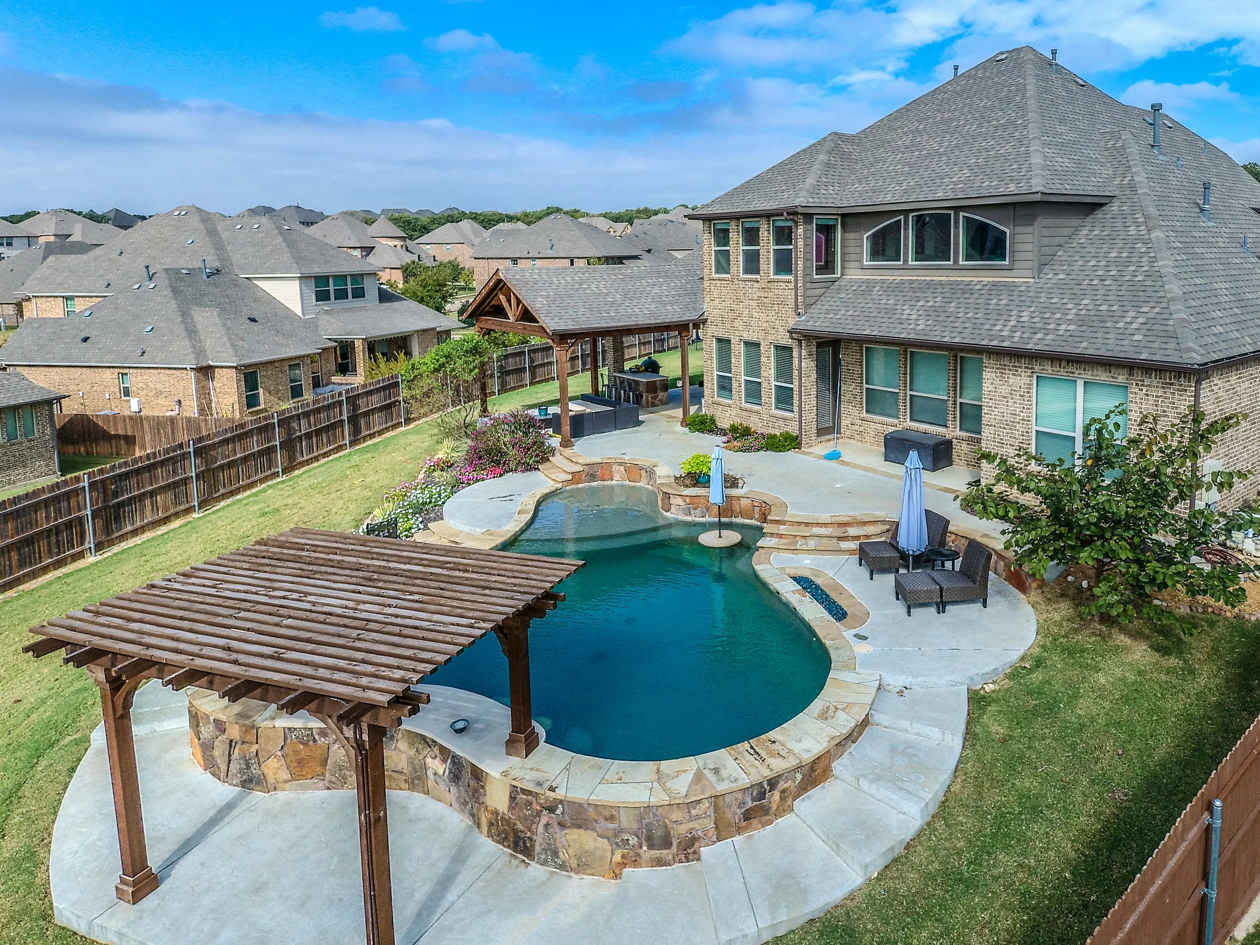 Backyard of a large house with a swimming pool, outdoor patio furniture, garden, and a gazebo, surrounded by a wooden fence and neighboring houses.