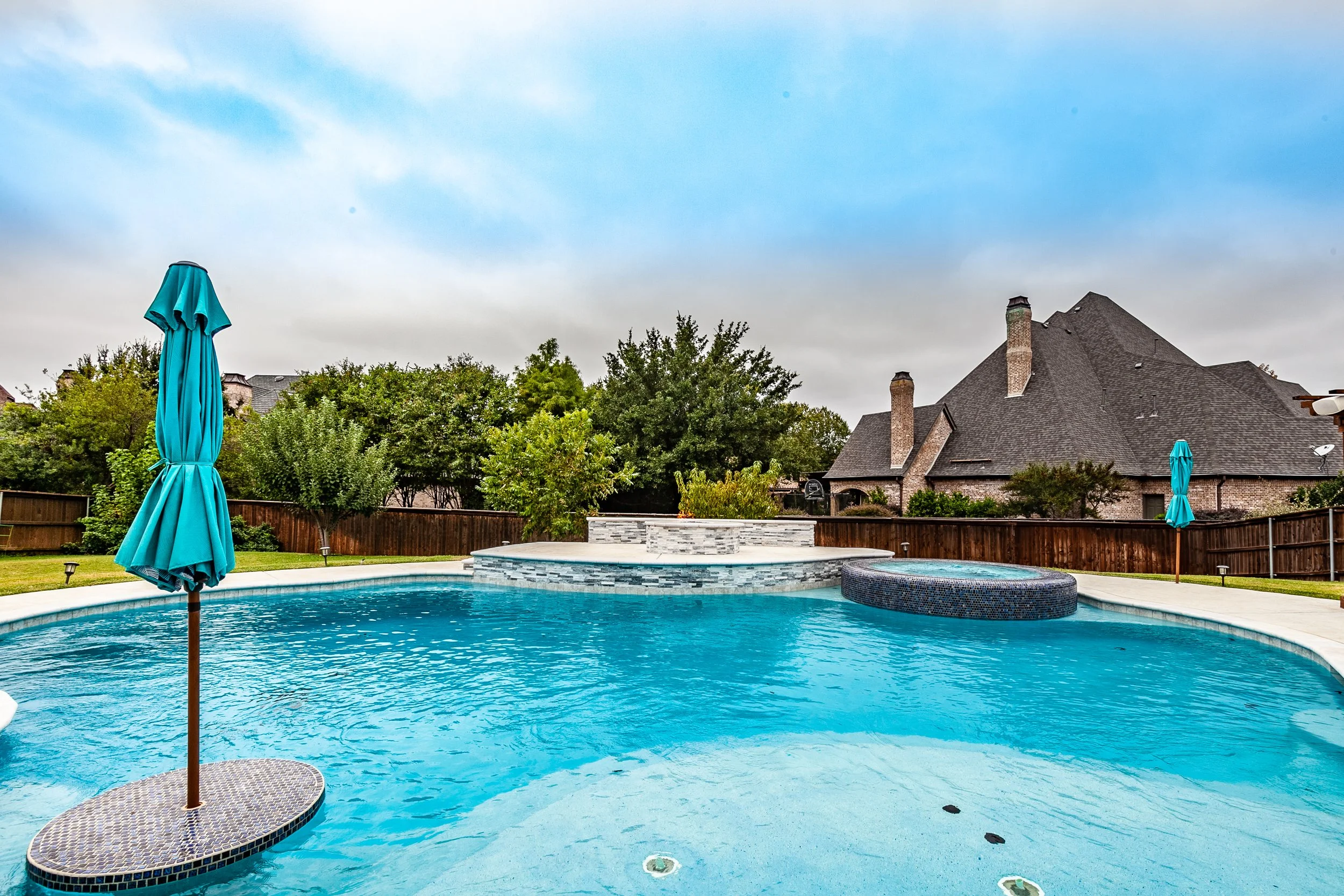 Backyard swimming pool with blue water, surrounded by a concrete deck, with two closed blue pool umbrellas, lush green trees, and a large brick house with a dark roof in the background, under a partly cloudy sky.