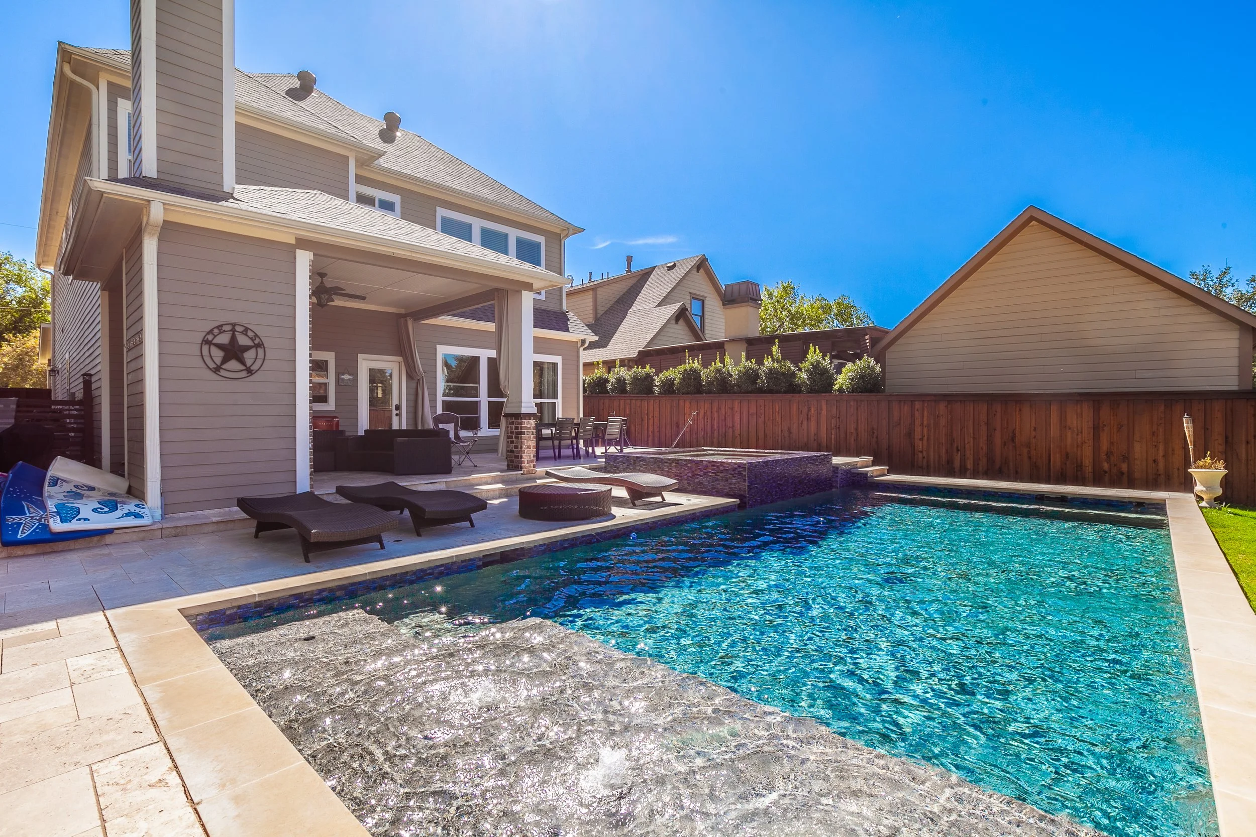 Backyard with a swimming pool, lounge chairs, and a patio with outdoor furniture in front of a house, with a wooden fence and neighboring houses in the background.