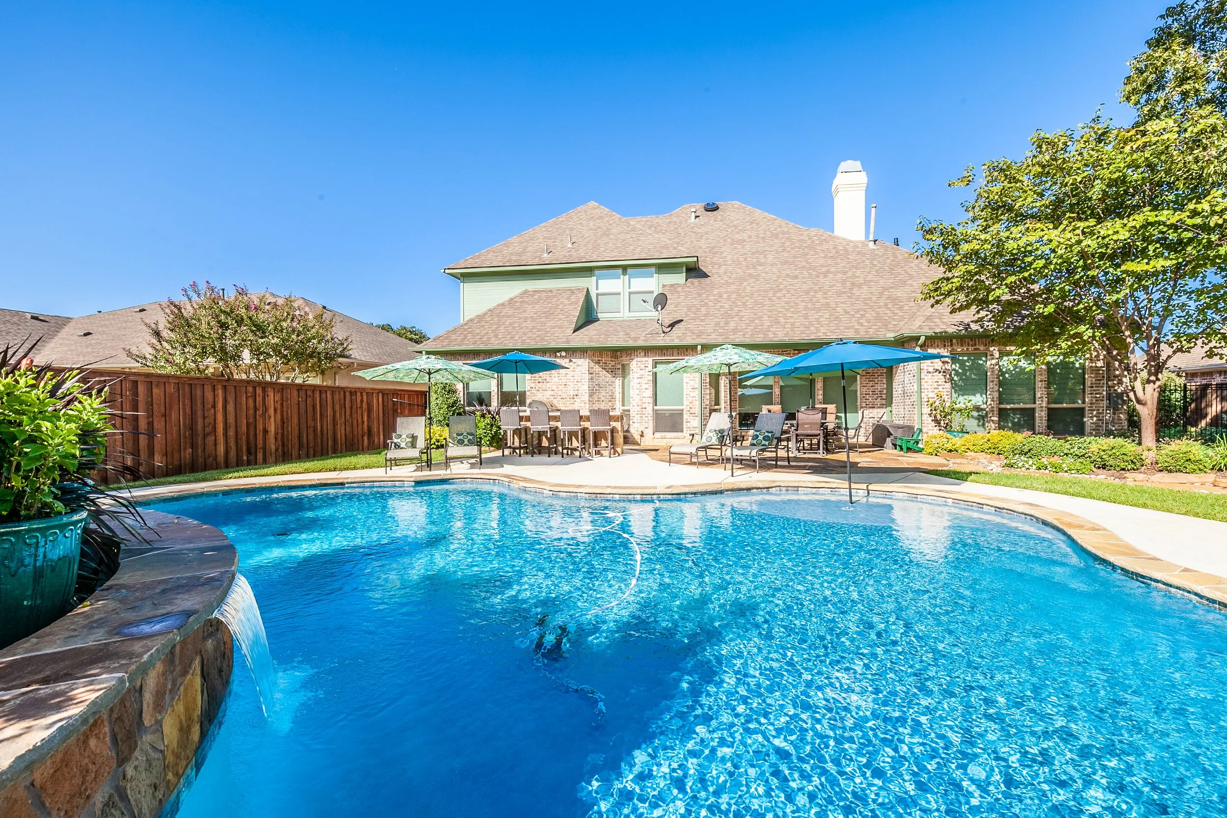 Backyard with a swimming pool, patio tables with umbrellas, and a house with a brick and green exterior, under a clear blue sky.