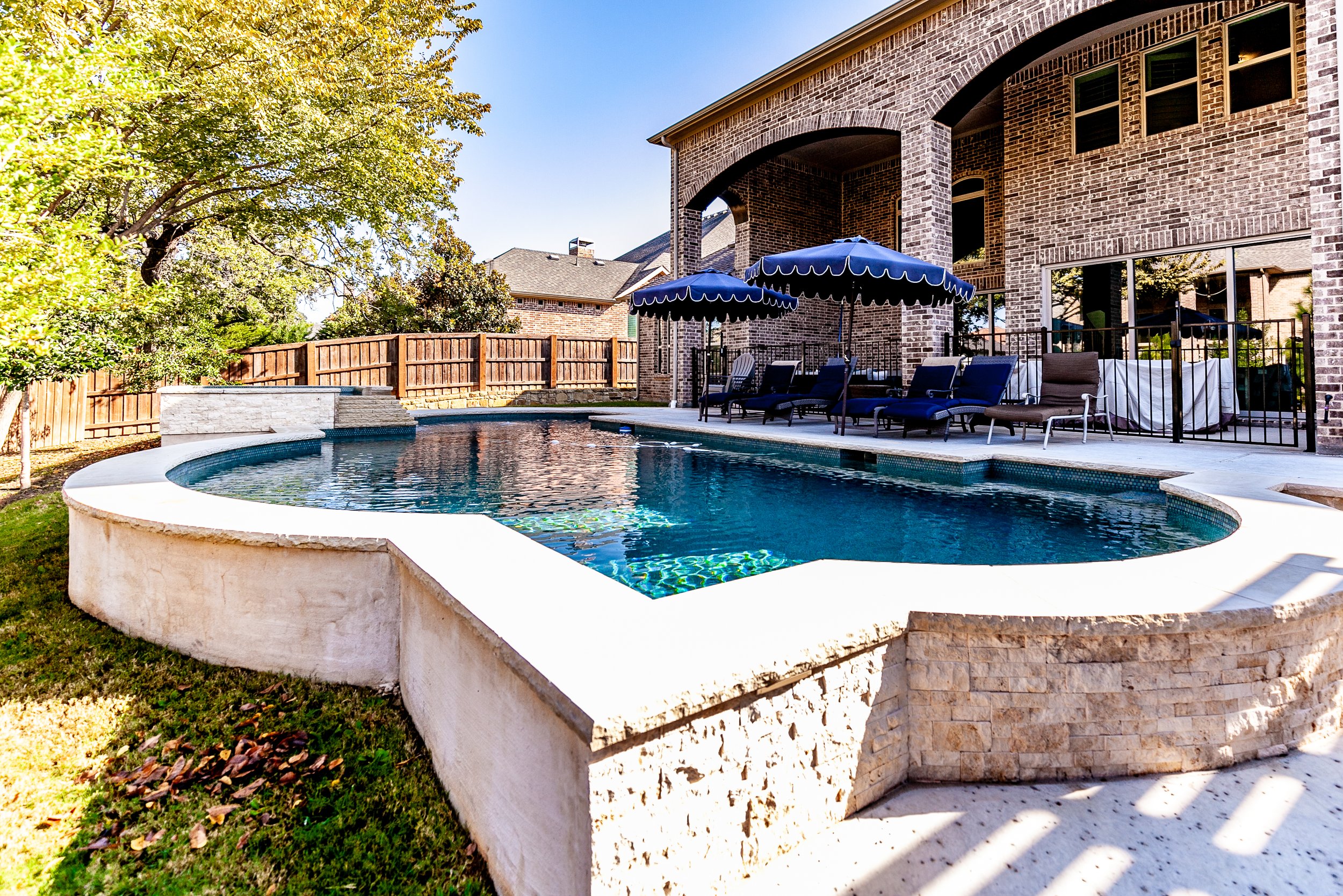 Backyard with a swimming pool, lounge chairs under umbrellas, and a house with a brick exterior in the background.