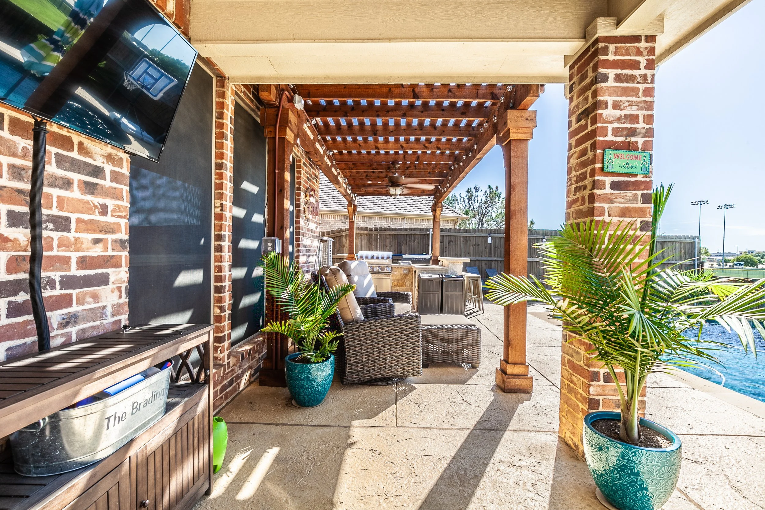 Outdoor patio with brick columns, wicker chairs, potted plants, a wall-mounted TV, and a view of a sports field in the distance.