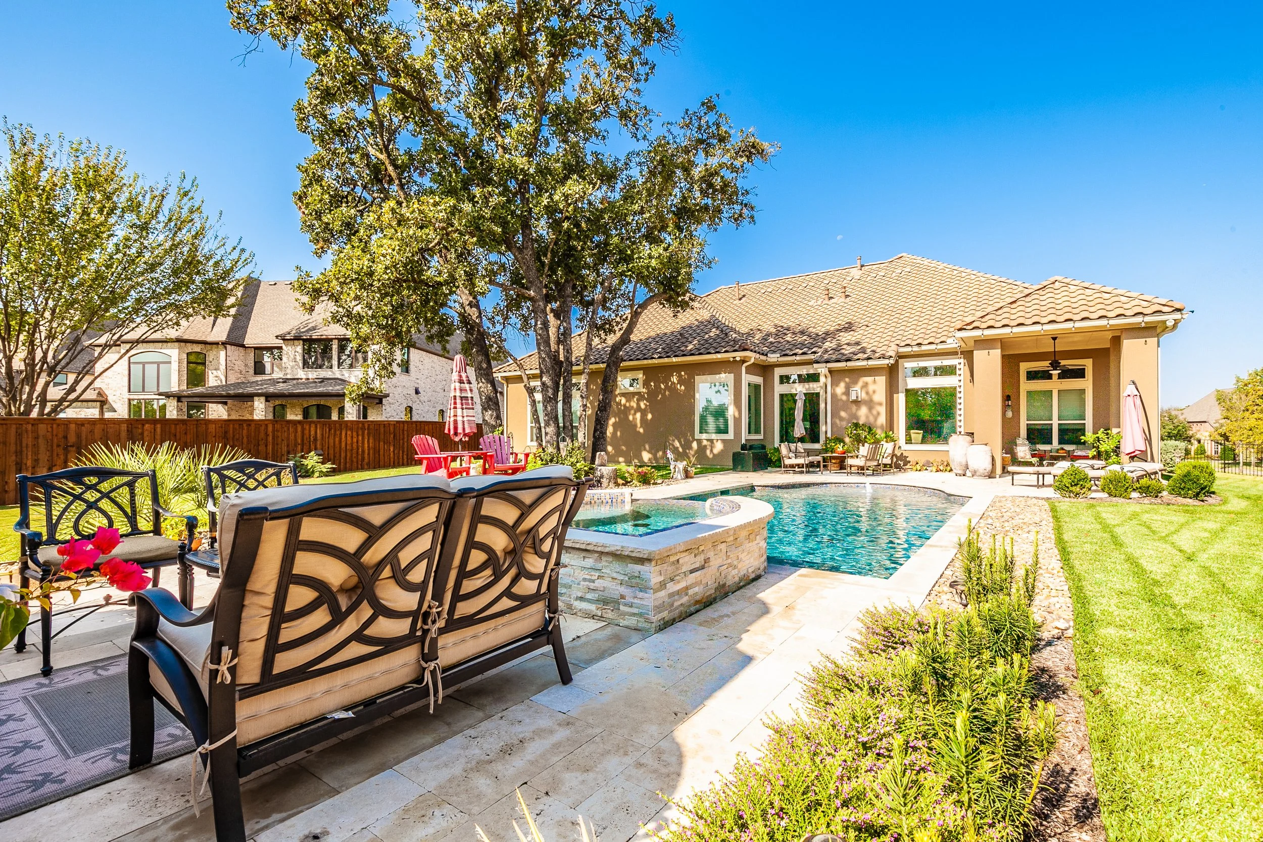 Backyard with swimming pool, patio furniture, trees, grass, and a house under a clear blue sky.