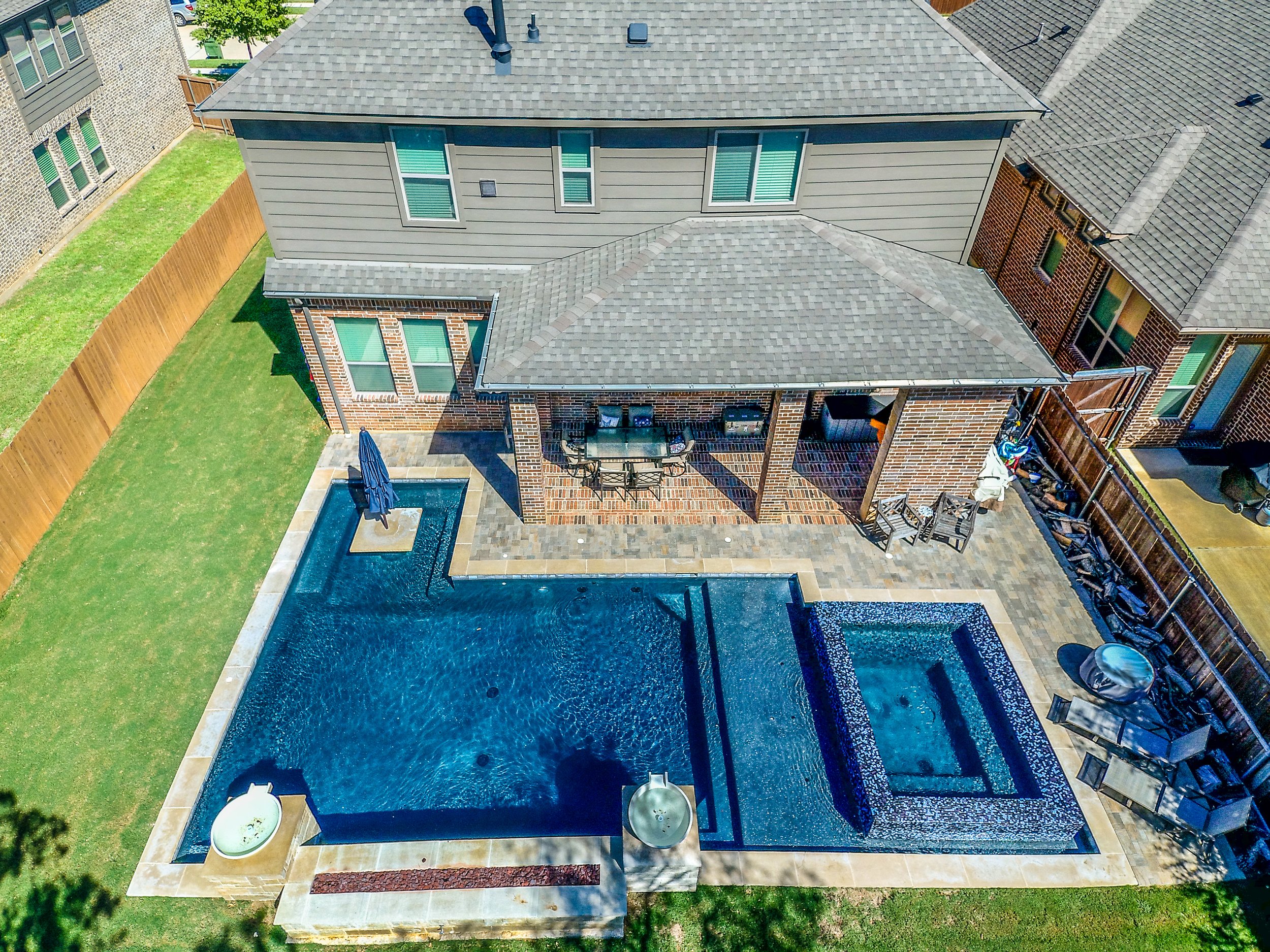 A backyard with a swimming pool, hot tub, patio furniture, and a covered patio next to a two-story house with gray siding and brick accents. The yard is fenced with a wooden fence.