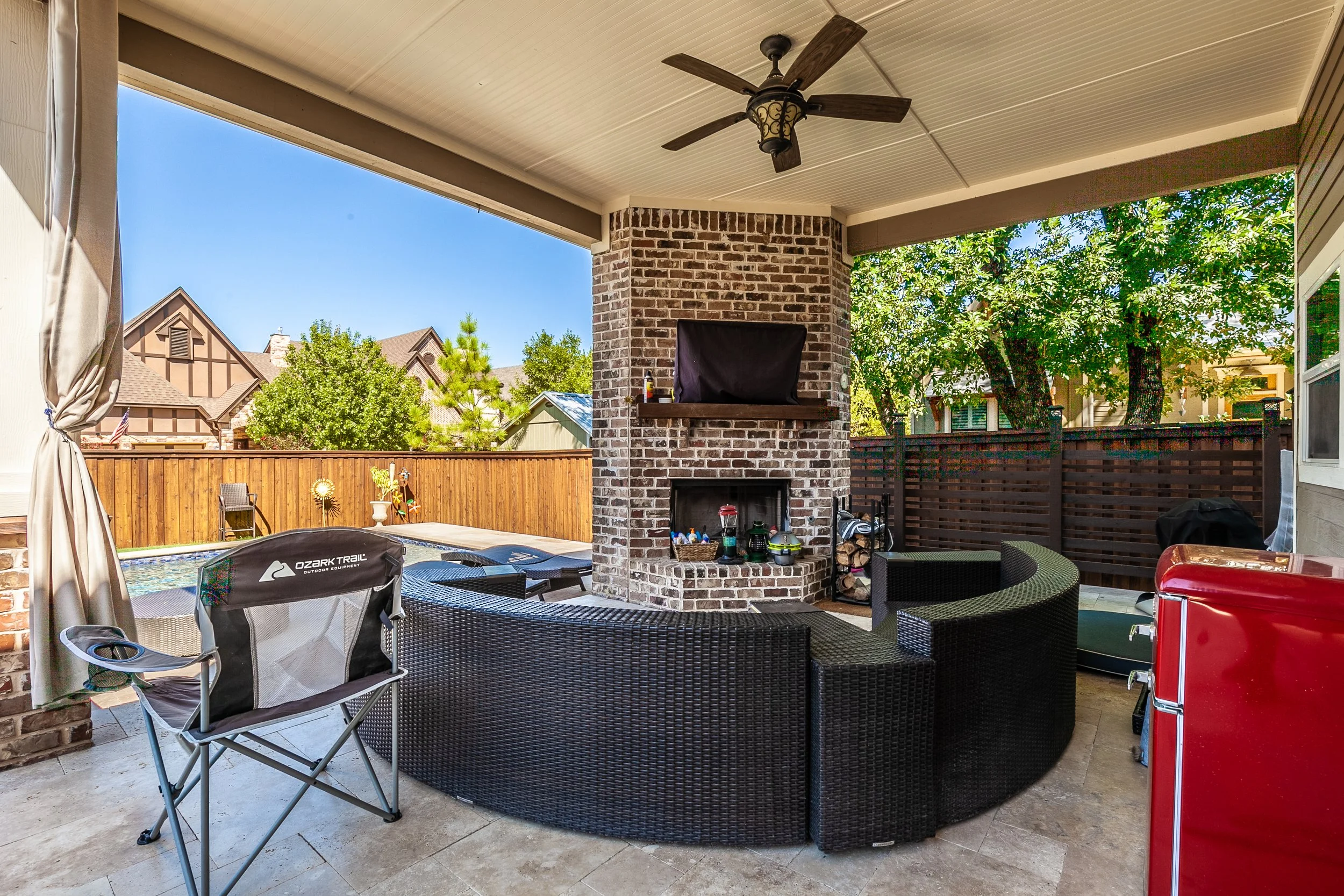 Covered backyard patio with outdoor furniture, a brick fireplace, a ceiling fan, and a view of a fenced yard with trees and neighboring houses.