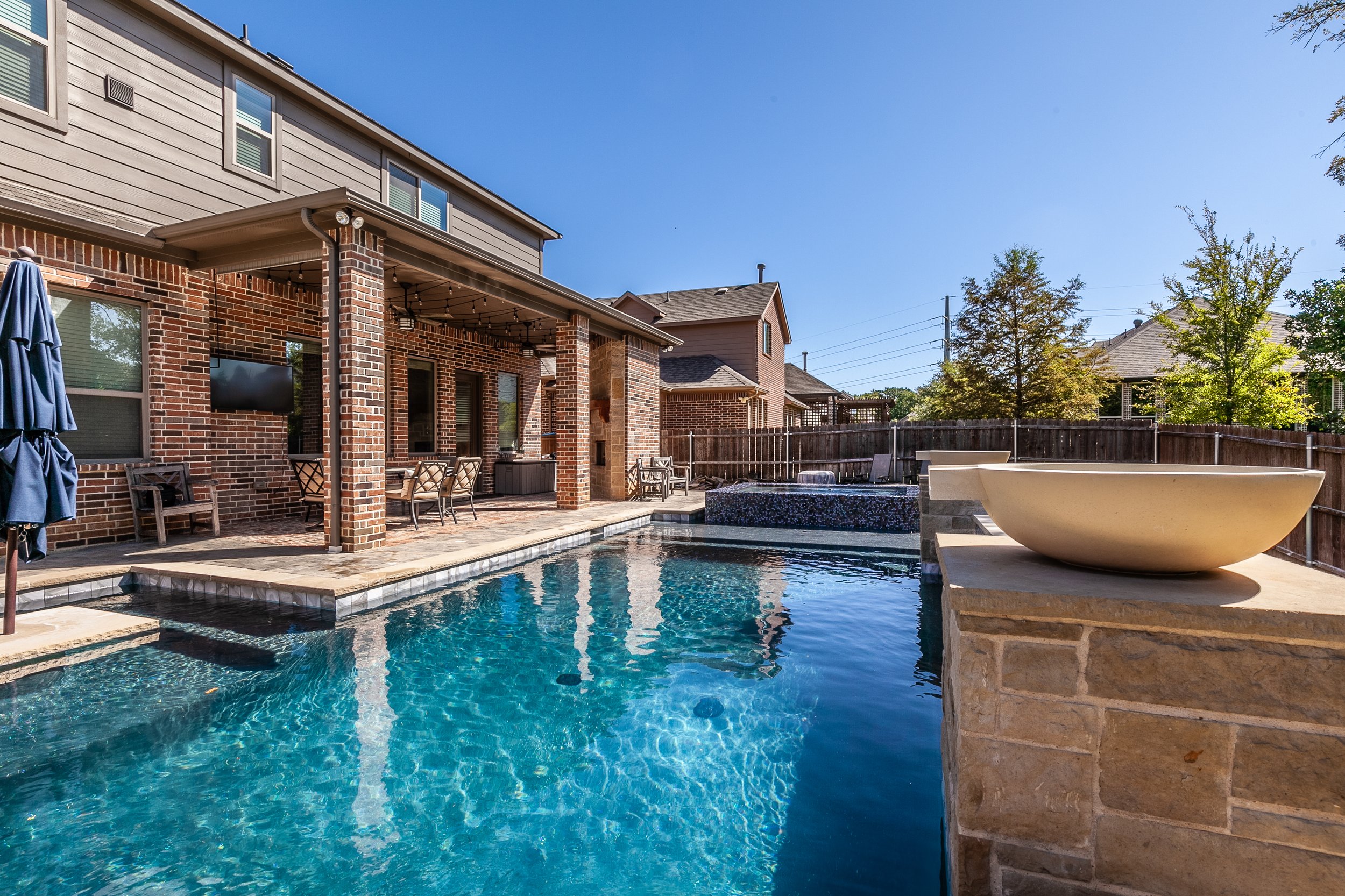 Backyard swimming pool area with a covered patio, tables, chairs, and a hot tub, surrounded by a wooden fence and neighboring houses, under a clear blue sky.