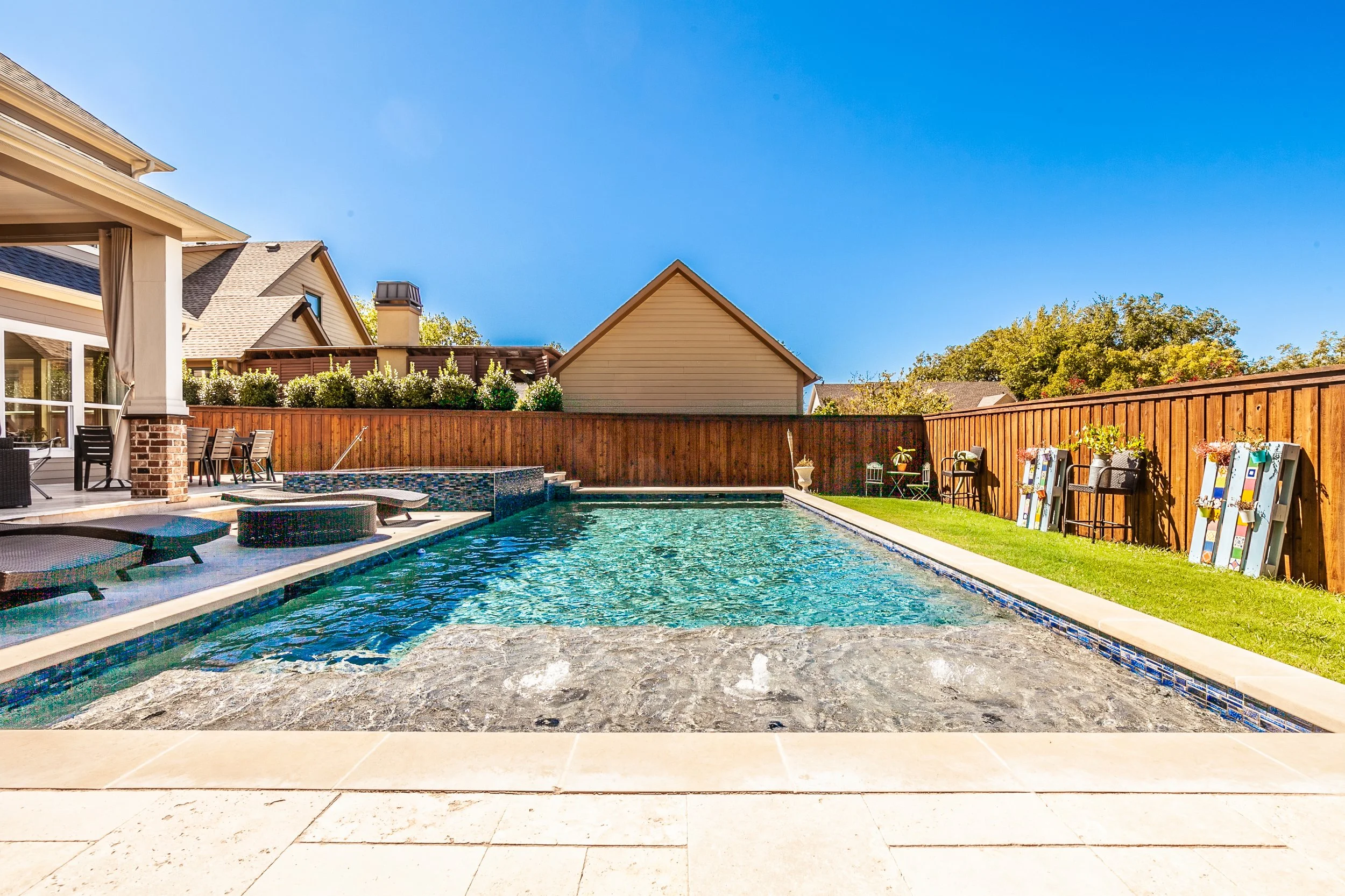 Bright backyard with a swimming pool, lounge chairs, a wooden fence, and surrounding greenery on a clear sunny day.