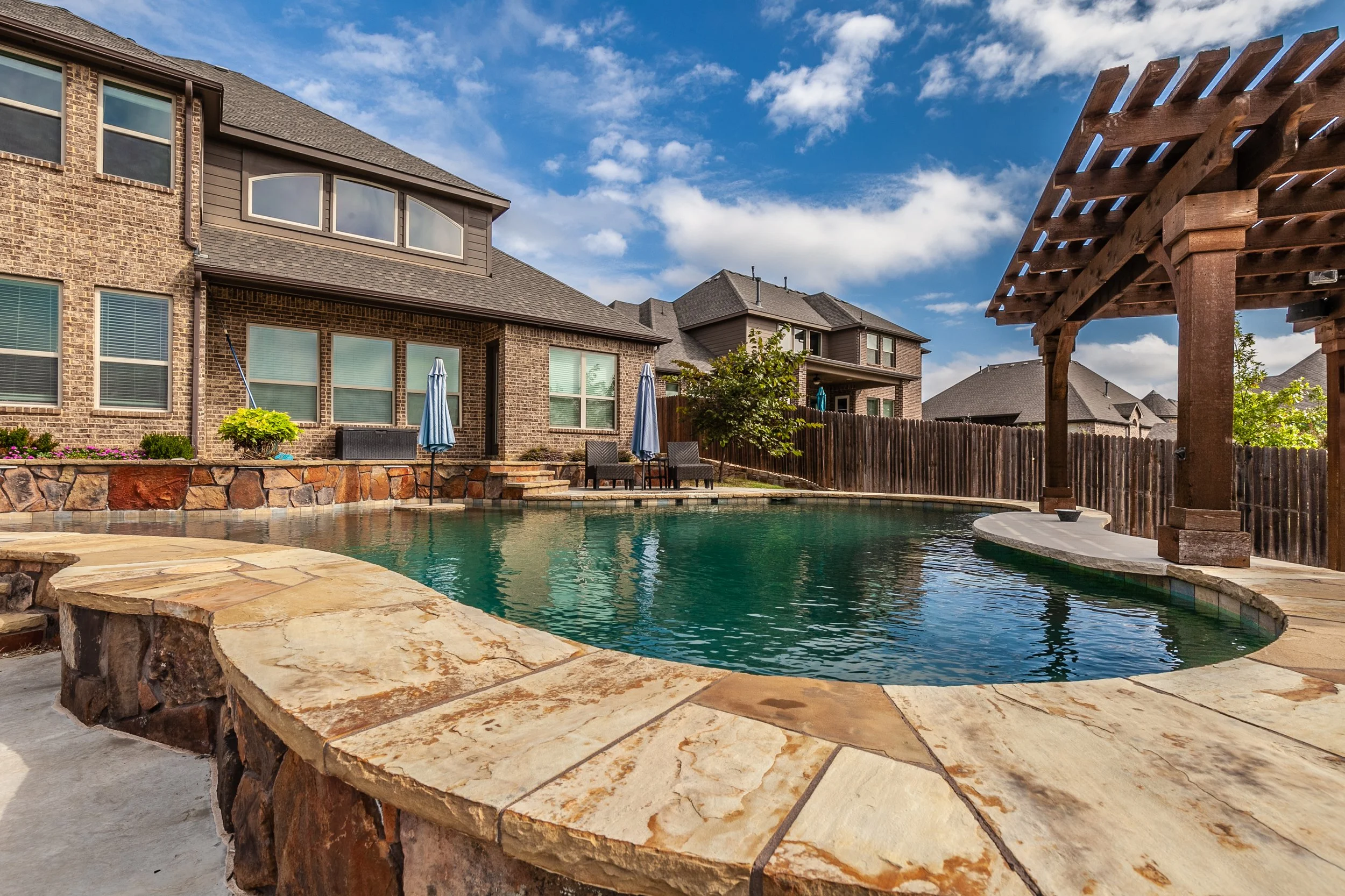 Residential backyard with a swimming pool, patio furniture, umbrellas, a wooden pergola, trees, and houses in the background under a partly cloudy sky.