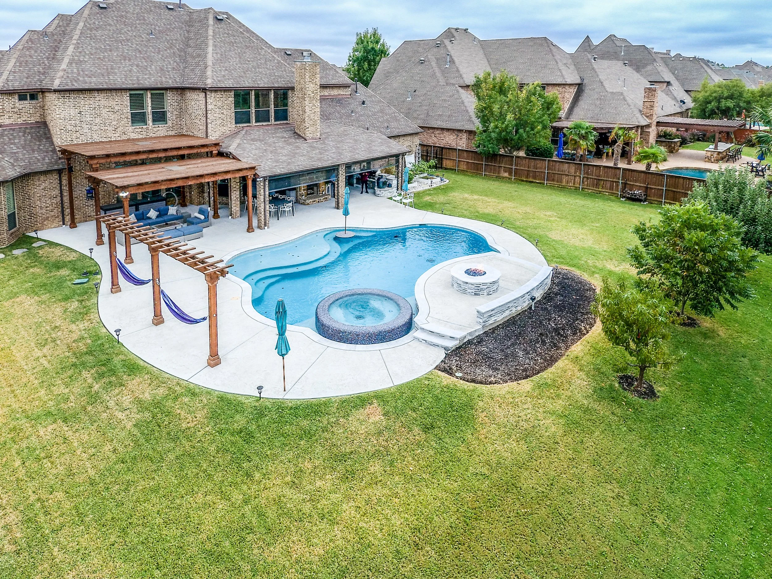 A backyard with a swimming pool, hot tub, fire pit, and a covered patio area with outdoor furniture, surrounded by a well-maintained lawn and a wooden fence.