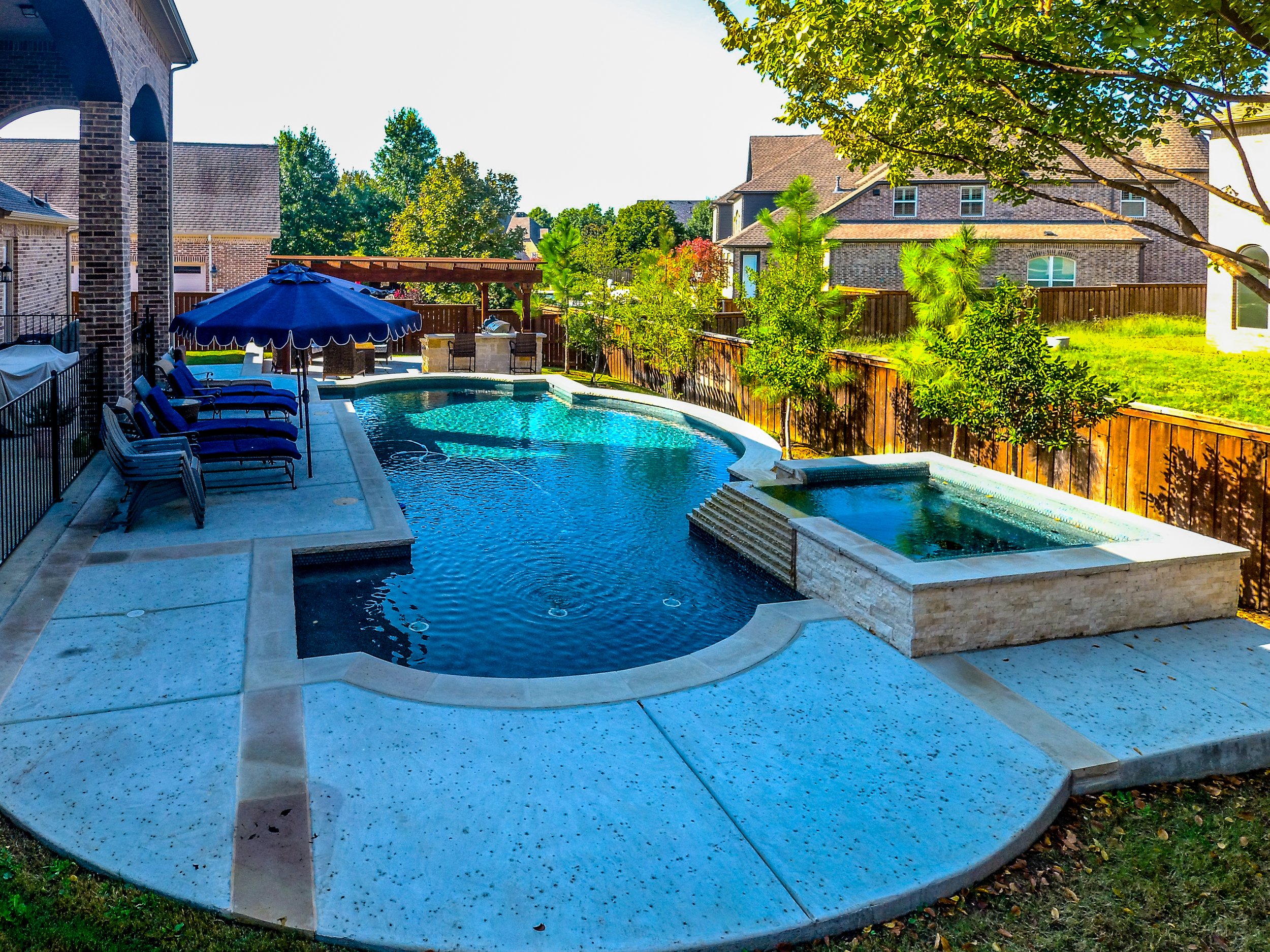 Backyard swimming pool with lounge chairs, umbrella, hot tub, and a shaded seating area, surrounded by a wooden fence and neighboring houses with trees and grassy lawns.