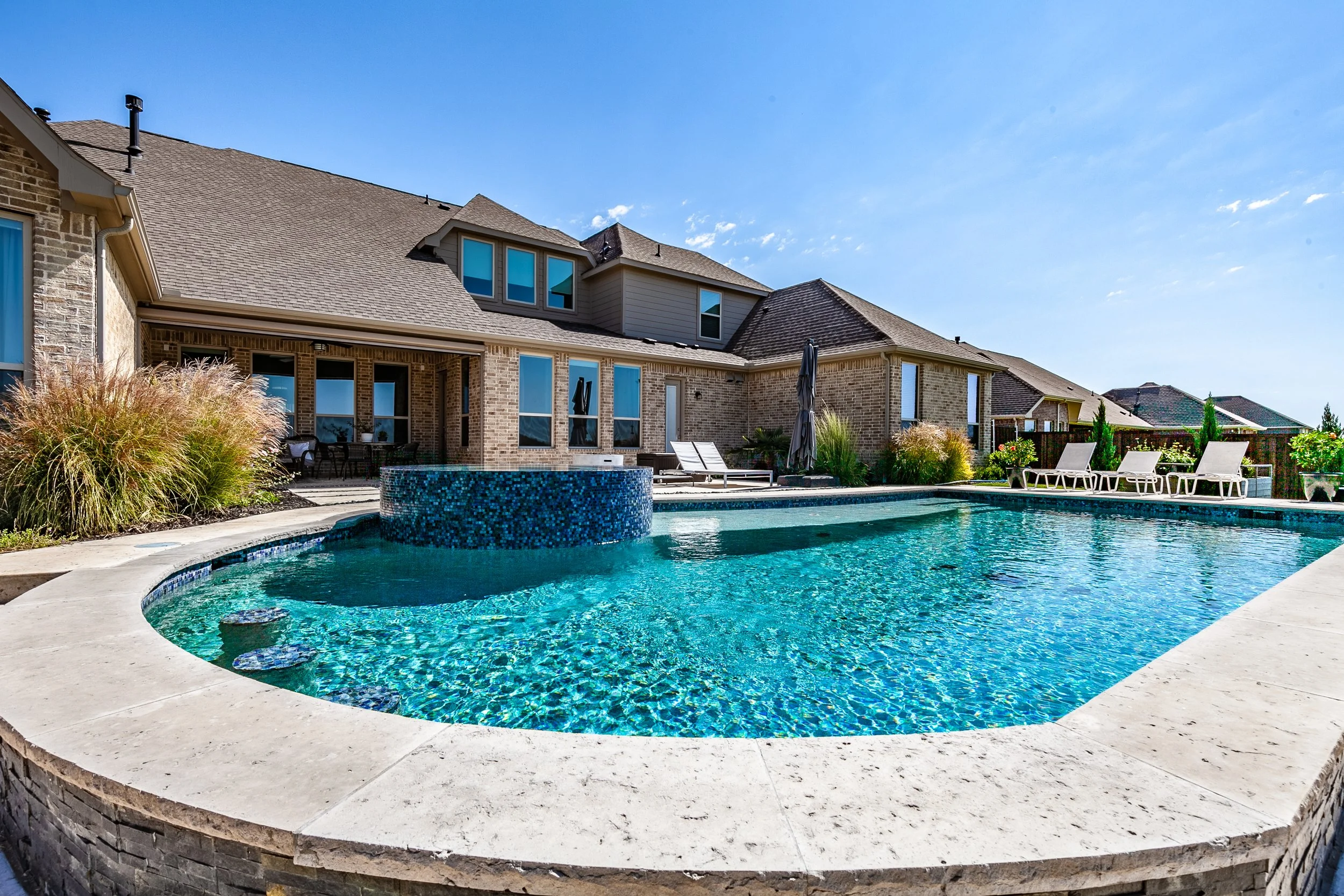 Backyard view of a house with a large swimming pool, lounge chairs, and landscaped plants under a blue sky.